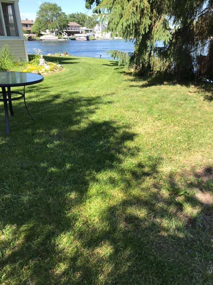 Shaded lakeside lawn with a patio table, trees, and houses across the water.