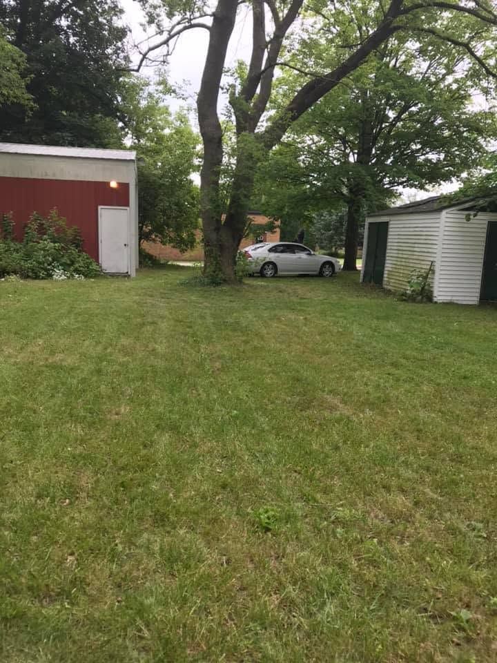 Grassy yard between a red shed and white garage, with a parked white car under trees.