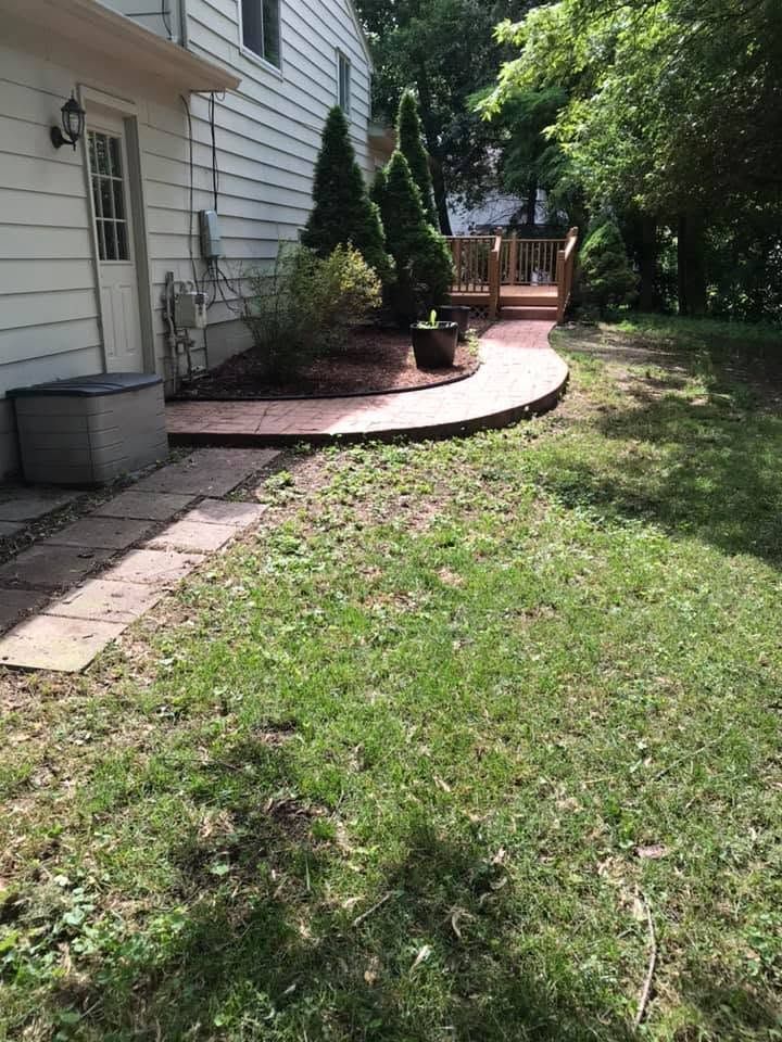 Curved brick garden path beside a white house, bordered by mulch and small shrubs, leading to a wooden gate.