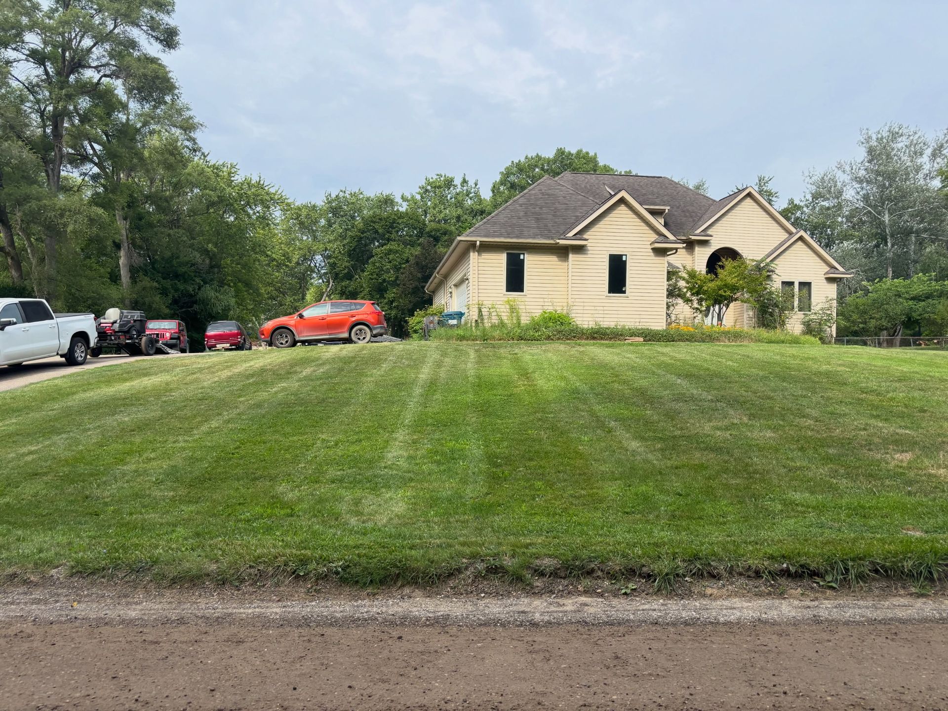 Suburban house with large mowed lawn, trees, and parked cars under a cloudy sky