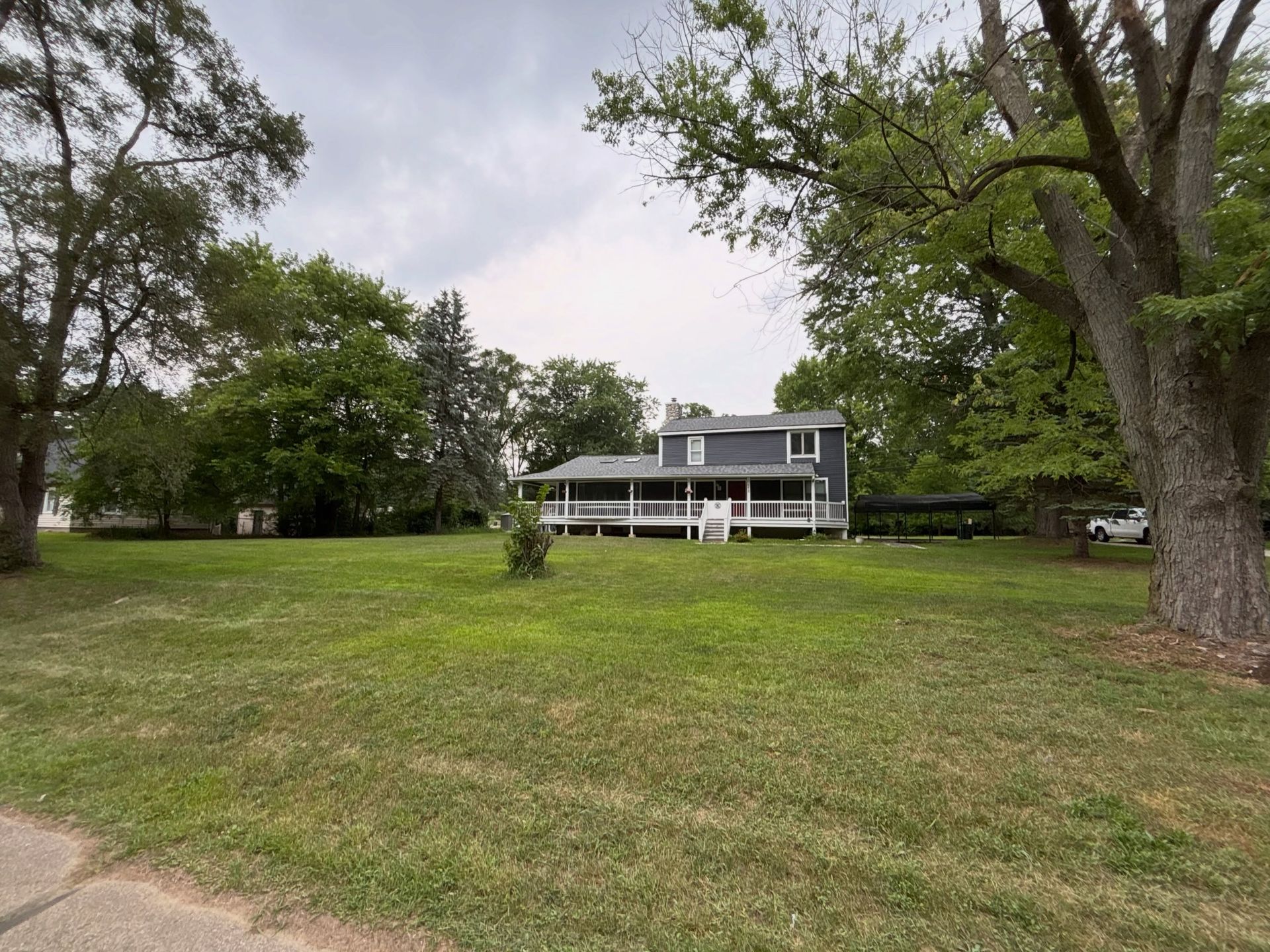 Large gray house with a white porch on a grassy lot, surrounded by tall trees under an overcast sky