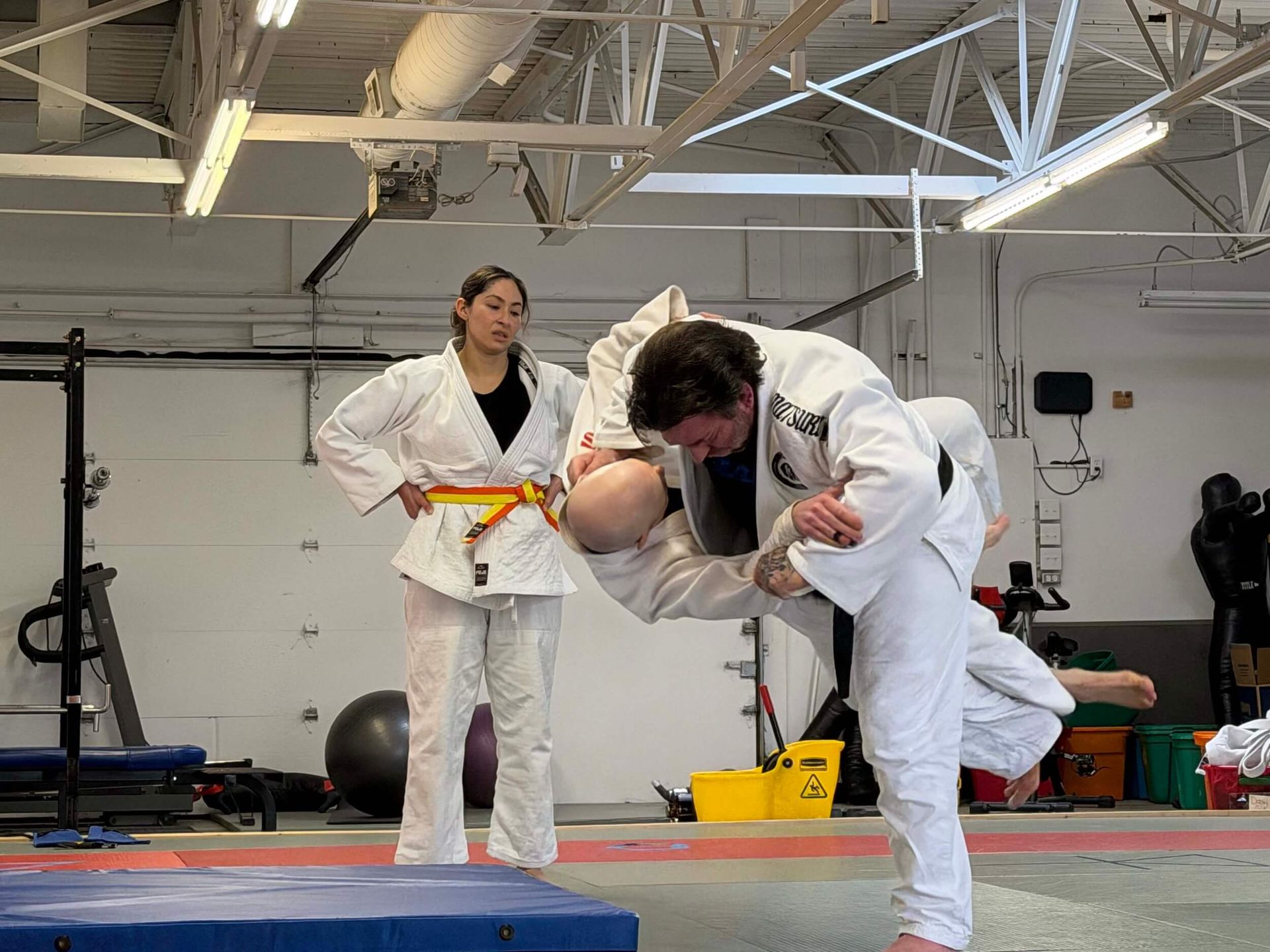 Two judo athletes in white gis practice a throw in a gym, with a coach watching nearby at Carmel Judo Indianapolis, IN