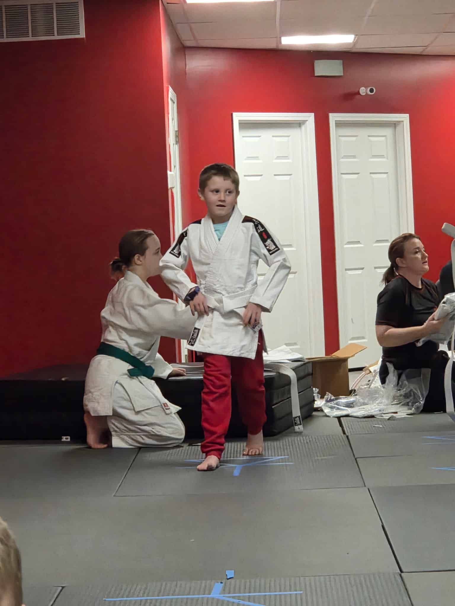Child in white martial arts uniform with red pants stands on a mat while others kneel in a red-walled dojo at Carmel Judo Indianapolis, IN