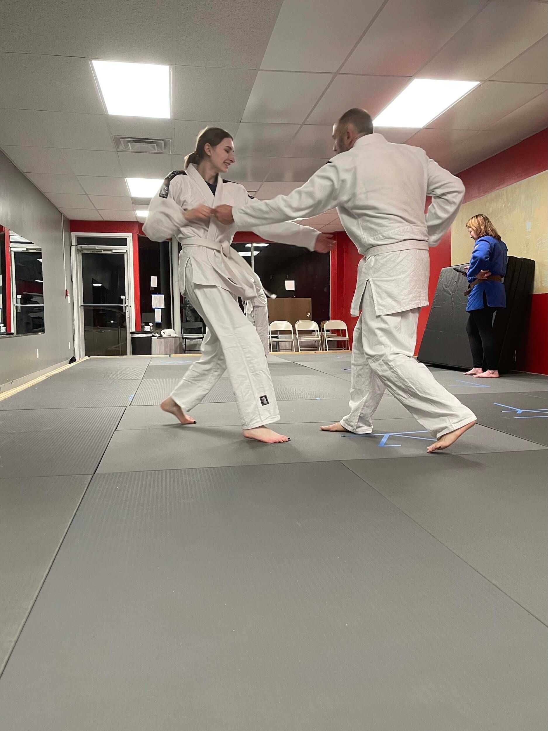 Two martial artists spar on padded mats in a studio, with a coach observing in the background at Carmel Judo Indianapolis, IN