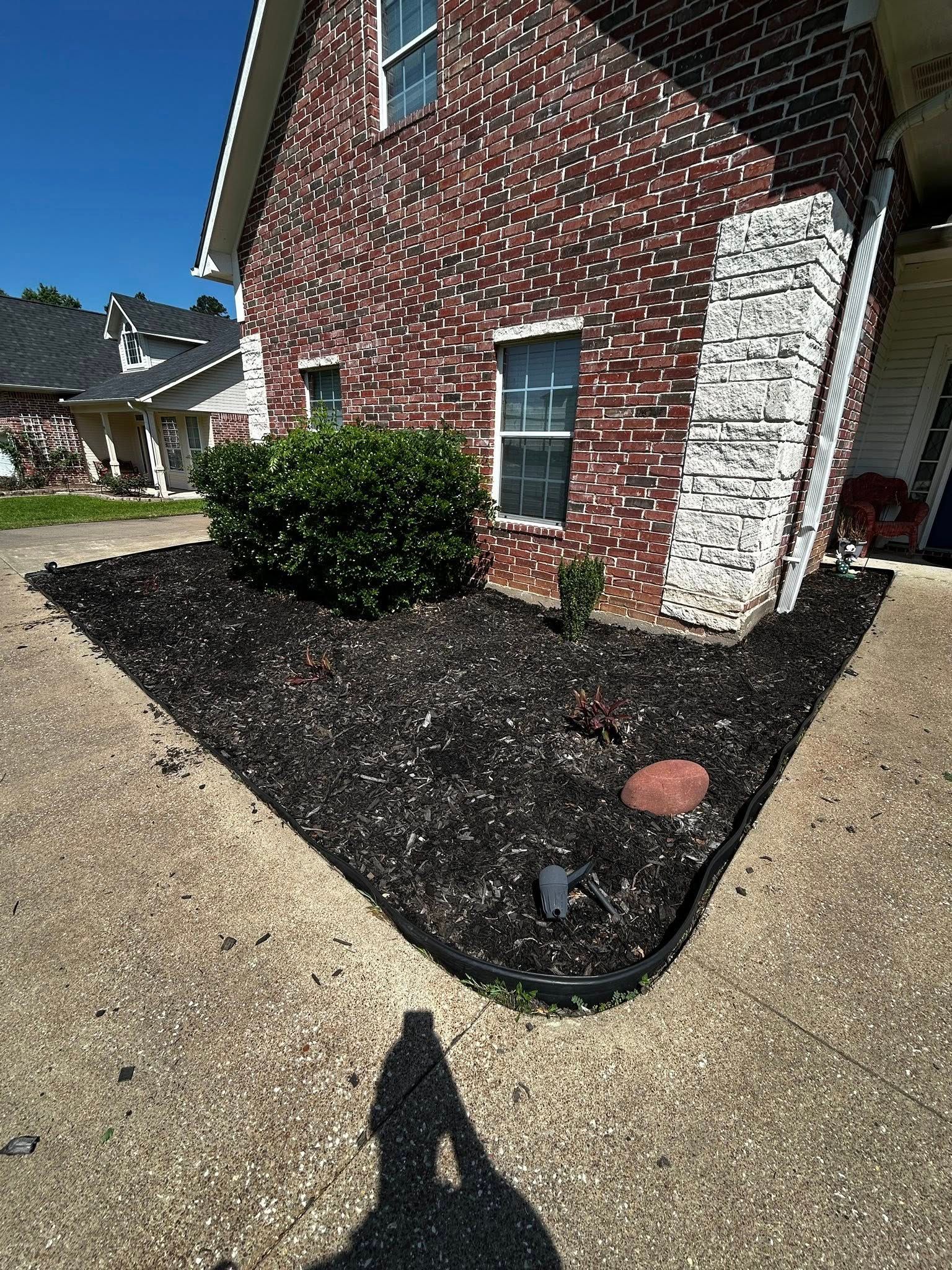 Brick house with black mulch flower bed in front. Concrete driveway, chimney on right, clear blue sky.