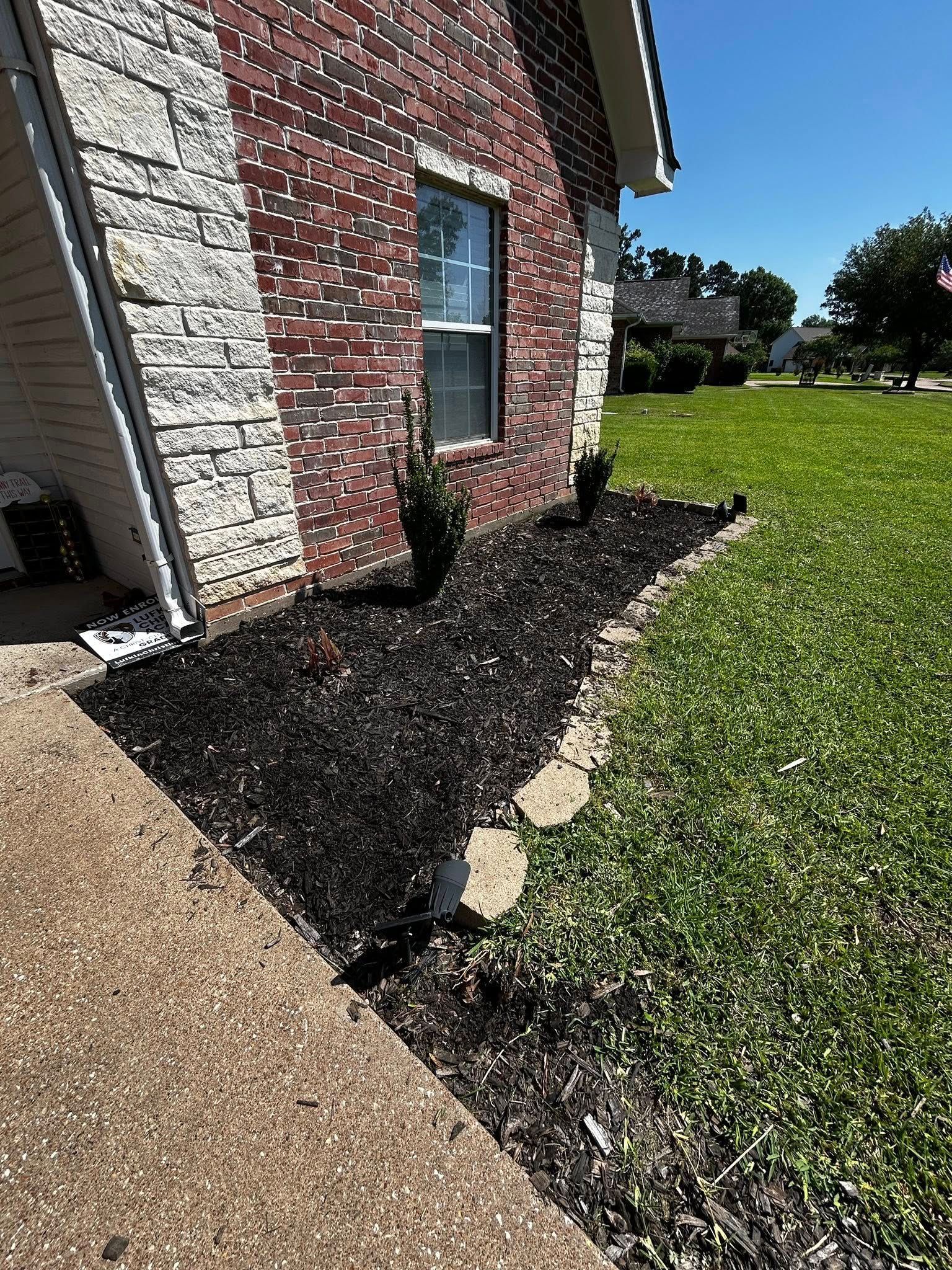 Red brick house with a flower bed lined with rocks and black mulch, and a green lawn.
