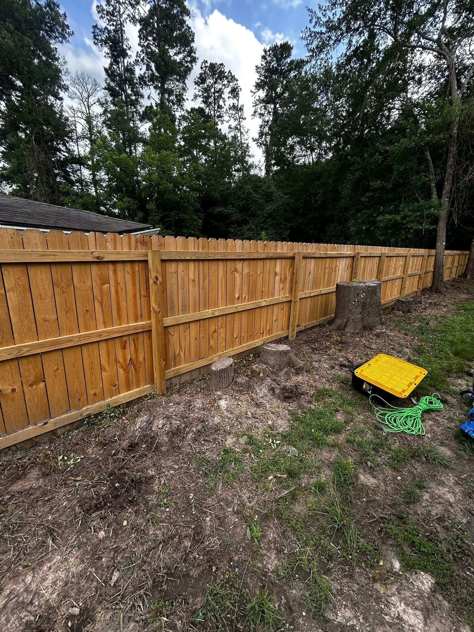 Wooden fence in a backyard with gravel and grass; trees in the background. A yellow and green item is on the grass.