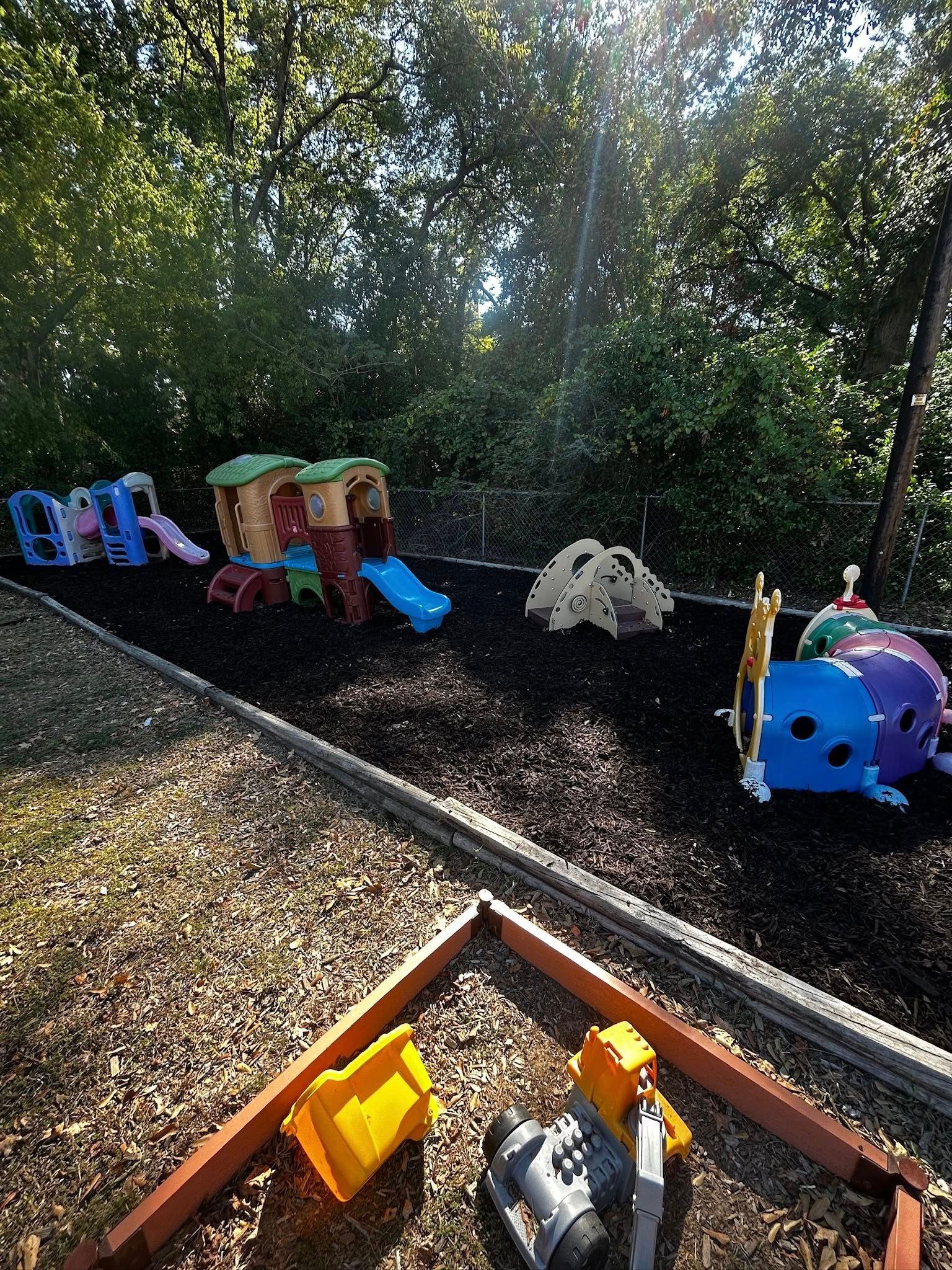 Playground with slides, playhouse, and toys on a black mulch surface, framed by wood and trees.