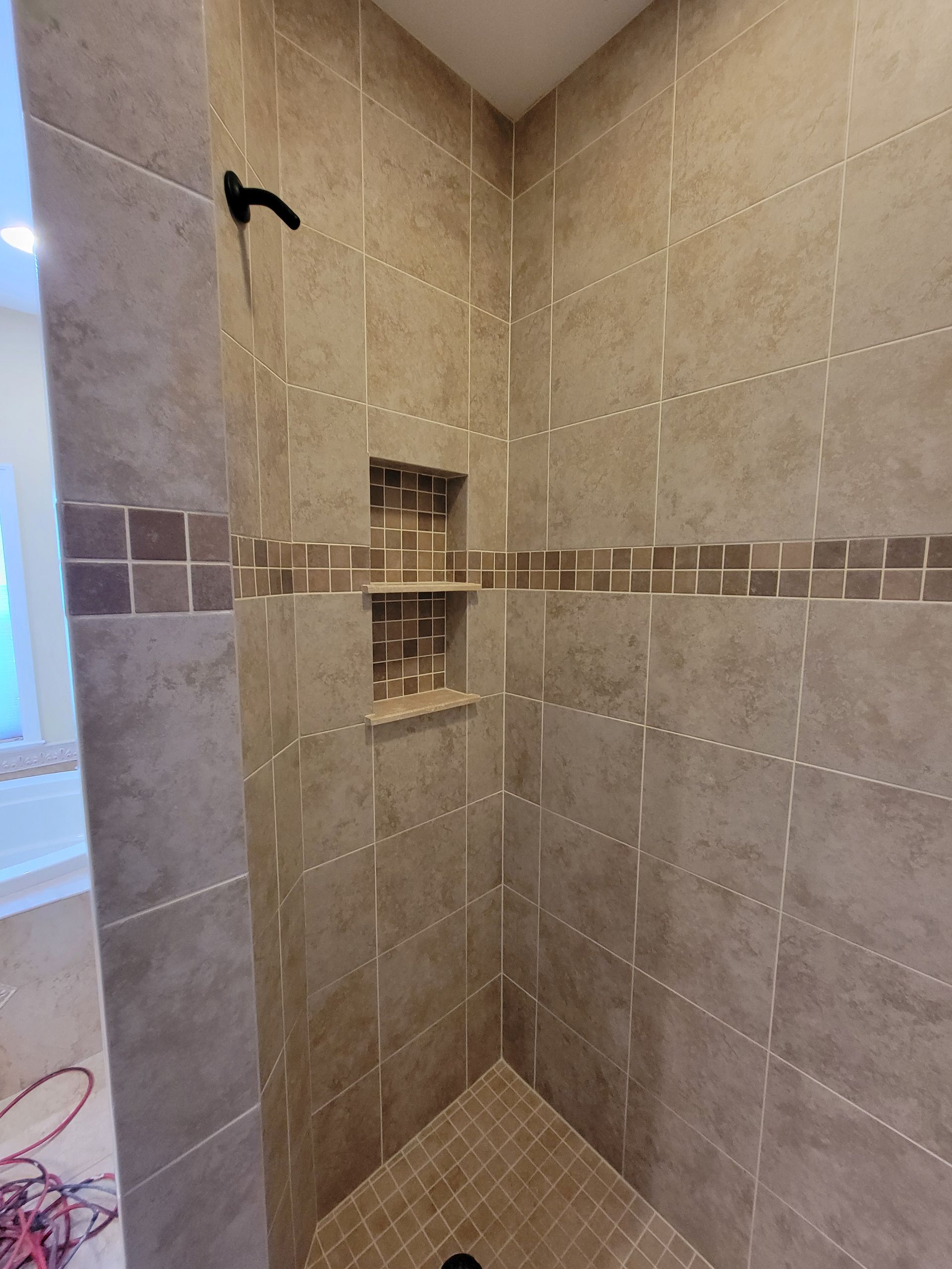 Tiled shower with corner seat. Beige tiles with a darker stripe. Two recessed shelves. Black shower head. Tiled shower with corner seat. Beige tiles with a darker stripe. Two recessed shelves. Black shower head.