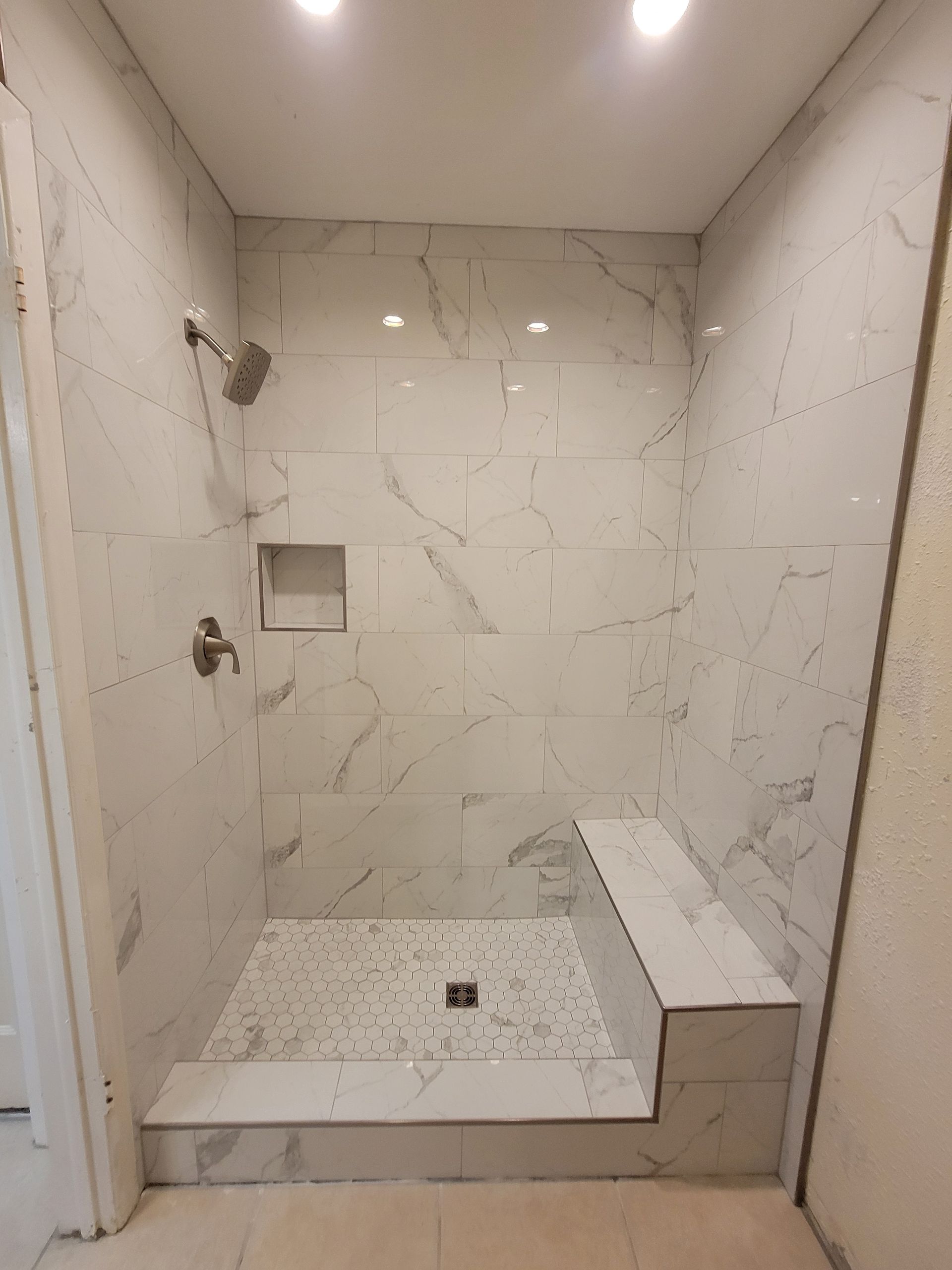Tiled shower stall with a built-in bench. White marble-look tiles, showerhead, and a recessed soap niche. Tiled shower stall with a built-in bench. White marble-look tiles, showerhead, and a recessed soap niche.