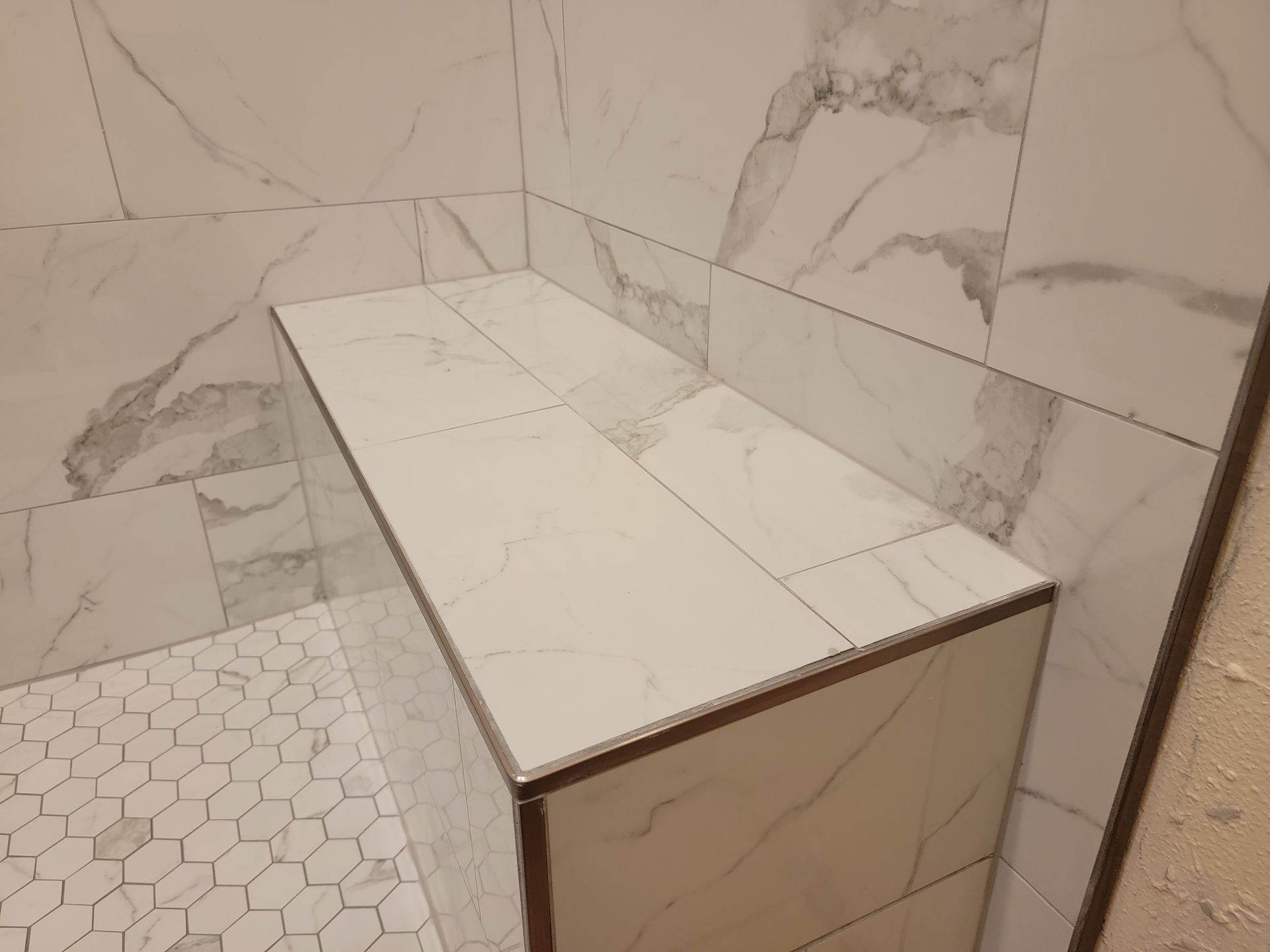 Tiled shower bench with white marble-patterned tiles; a corner shower. Hexagon tile floor. Tiled shower bench with white marble-patterned tiles; a corner shower. Hexagon tile floor.
