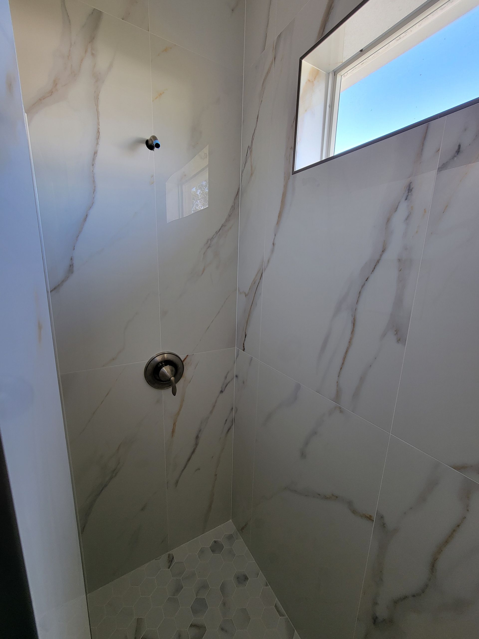 White tiled shower with marble-like veining, showerhead, faucet, and a small window with a blue sky visible.