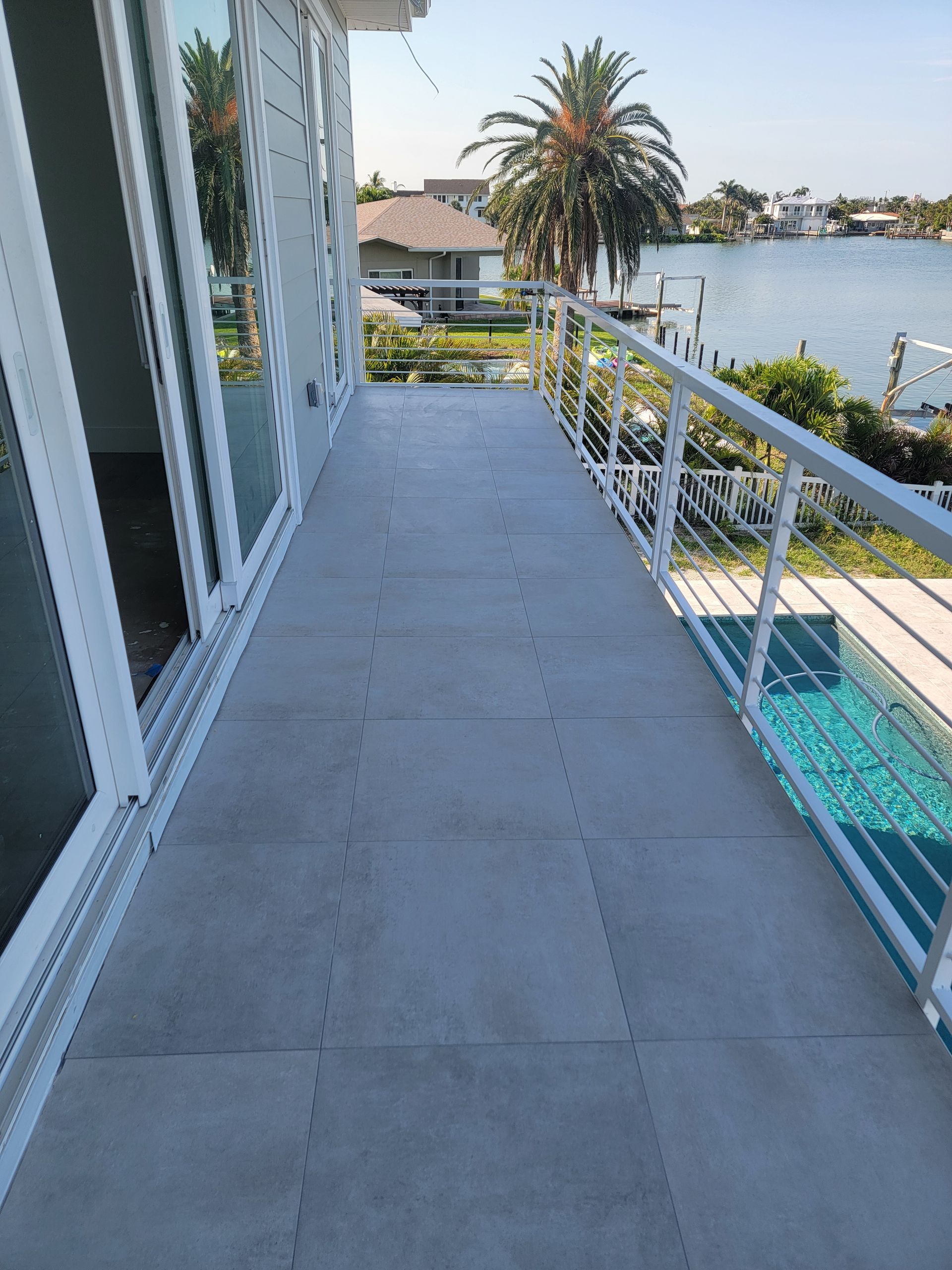 Balcony overlooking water, white railing, gray tiled floor, sliding glass doors, palm tree in background. Balcony overlooking water, white railing, gray tiled floor, sliding glass doors, palm tree in background.