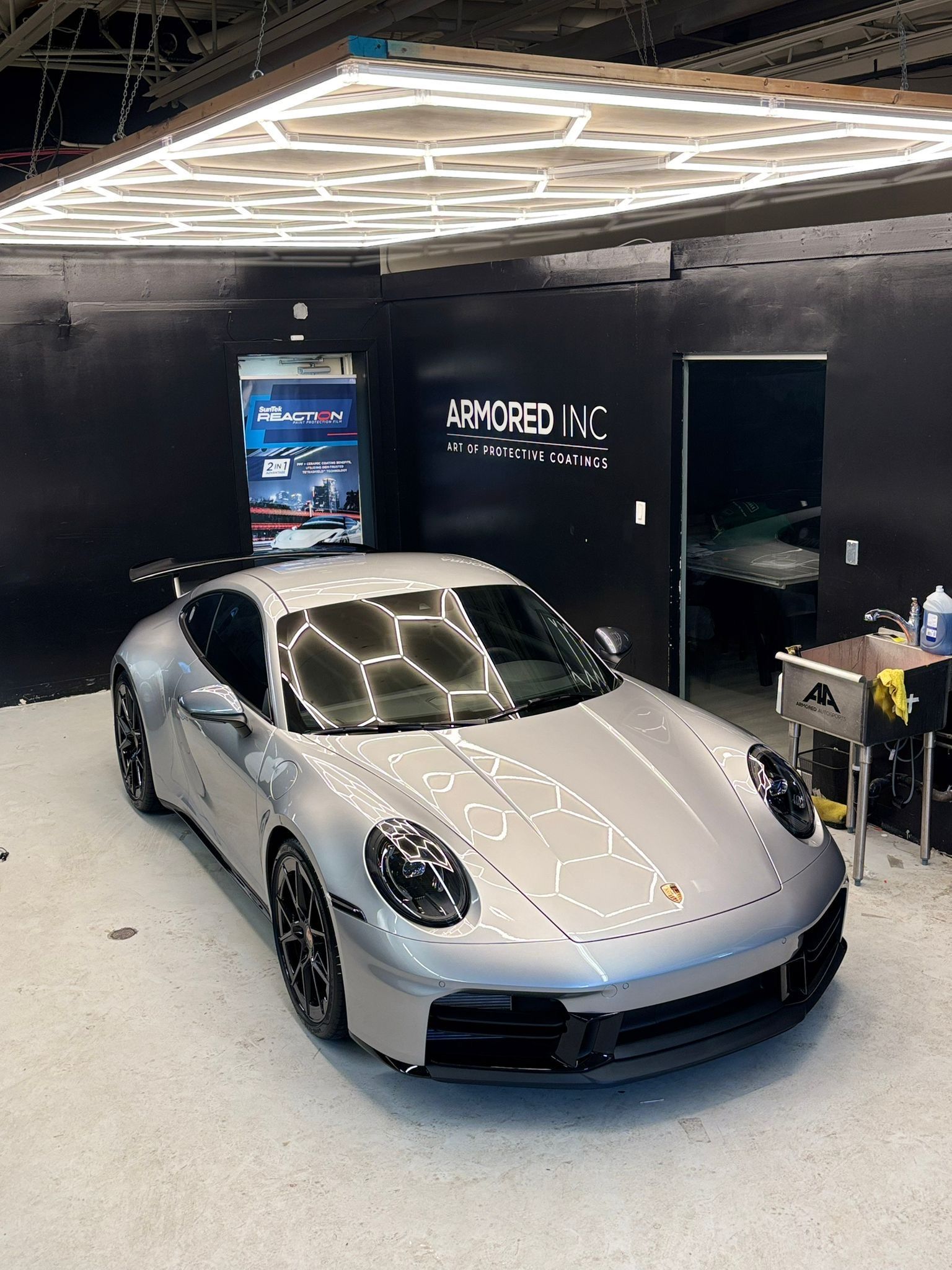 A silver Porsche sports car parked inside a professional automotive detailing shop with hexagon ceiling lighting.