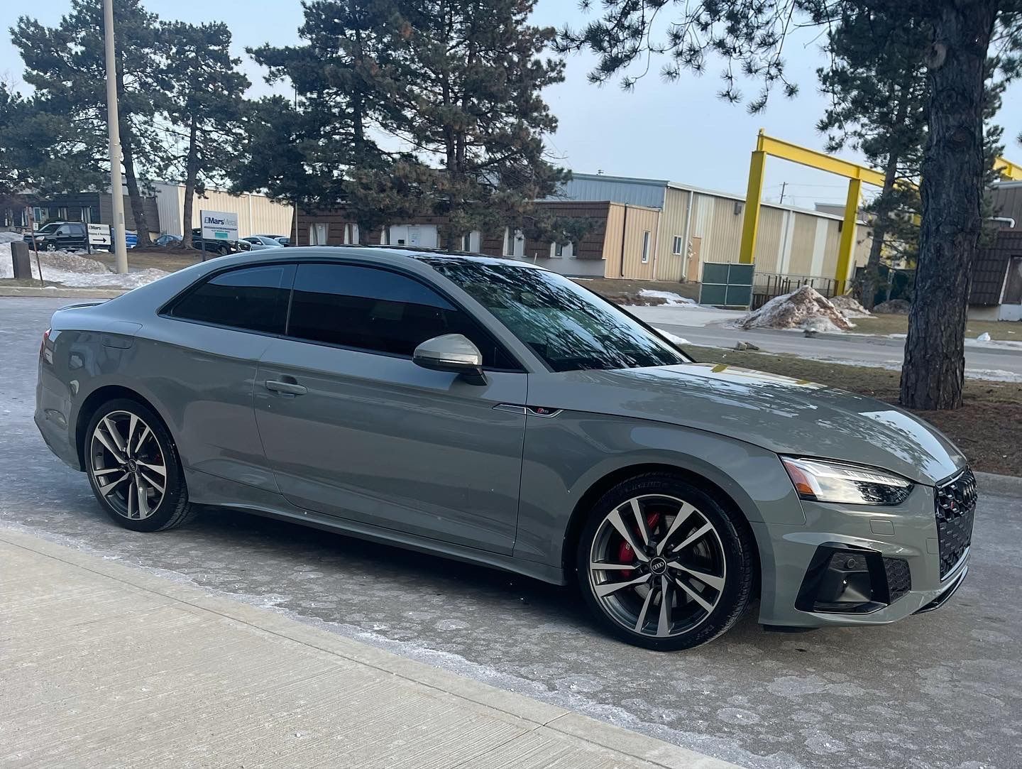 A gray Audi A5 coupe parked on a paved lot with light snow patches, near trees and a yellow industrial structure.