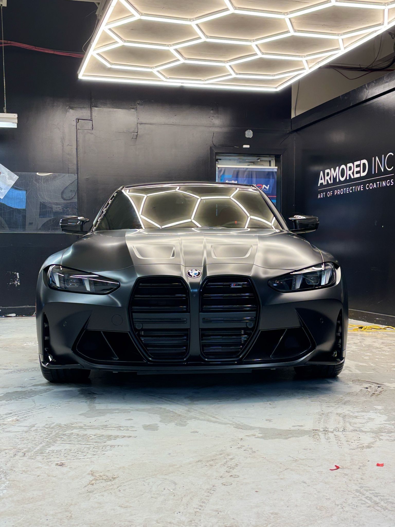 A matte black BMW M3 with its iconic vertical grille parked in a studio with a hexagonal LED ceiling light.