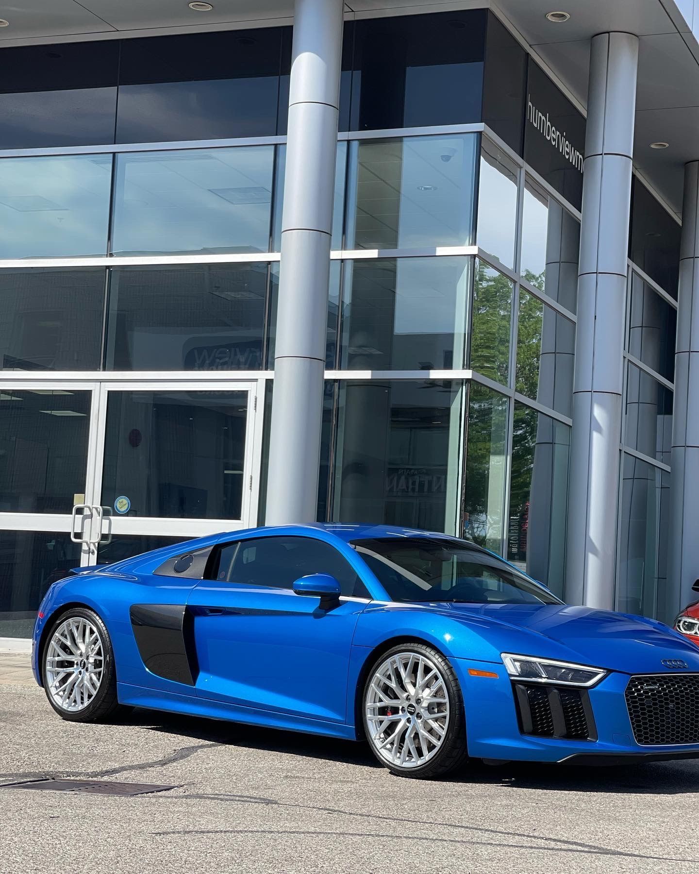 A vibrant blue Audi R8 sports car parked in front of a modern glass-fronted dealership building.
