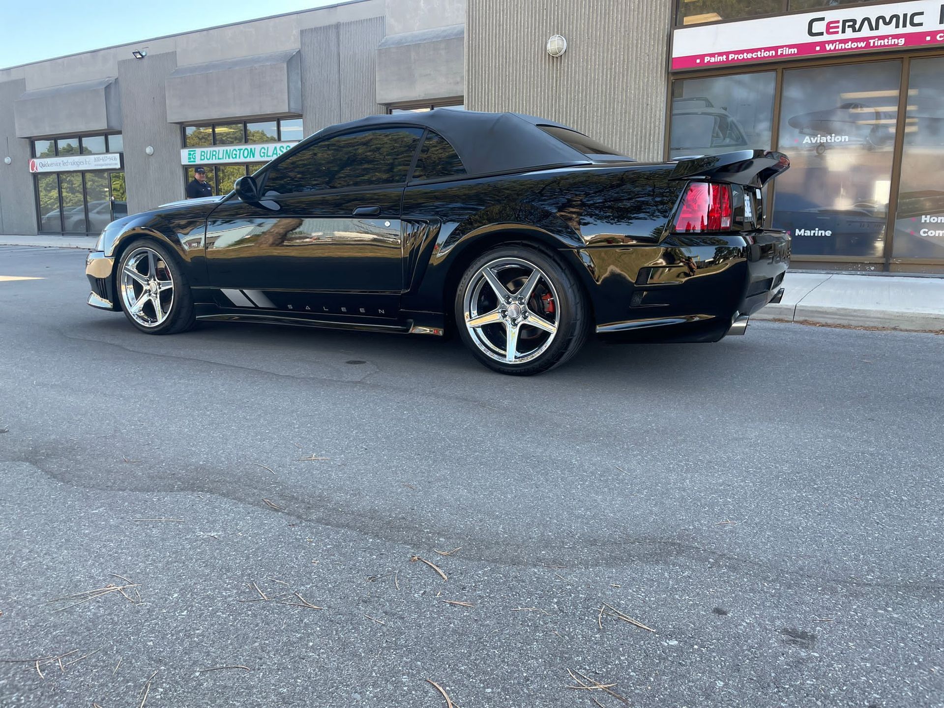 A sleek black convertible Ford Mustang parked on an asphalt street in front of a commercial storefront.