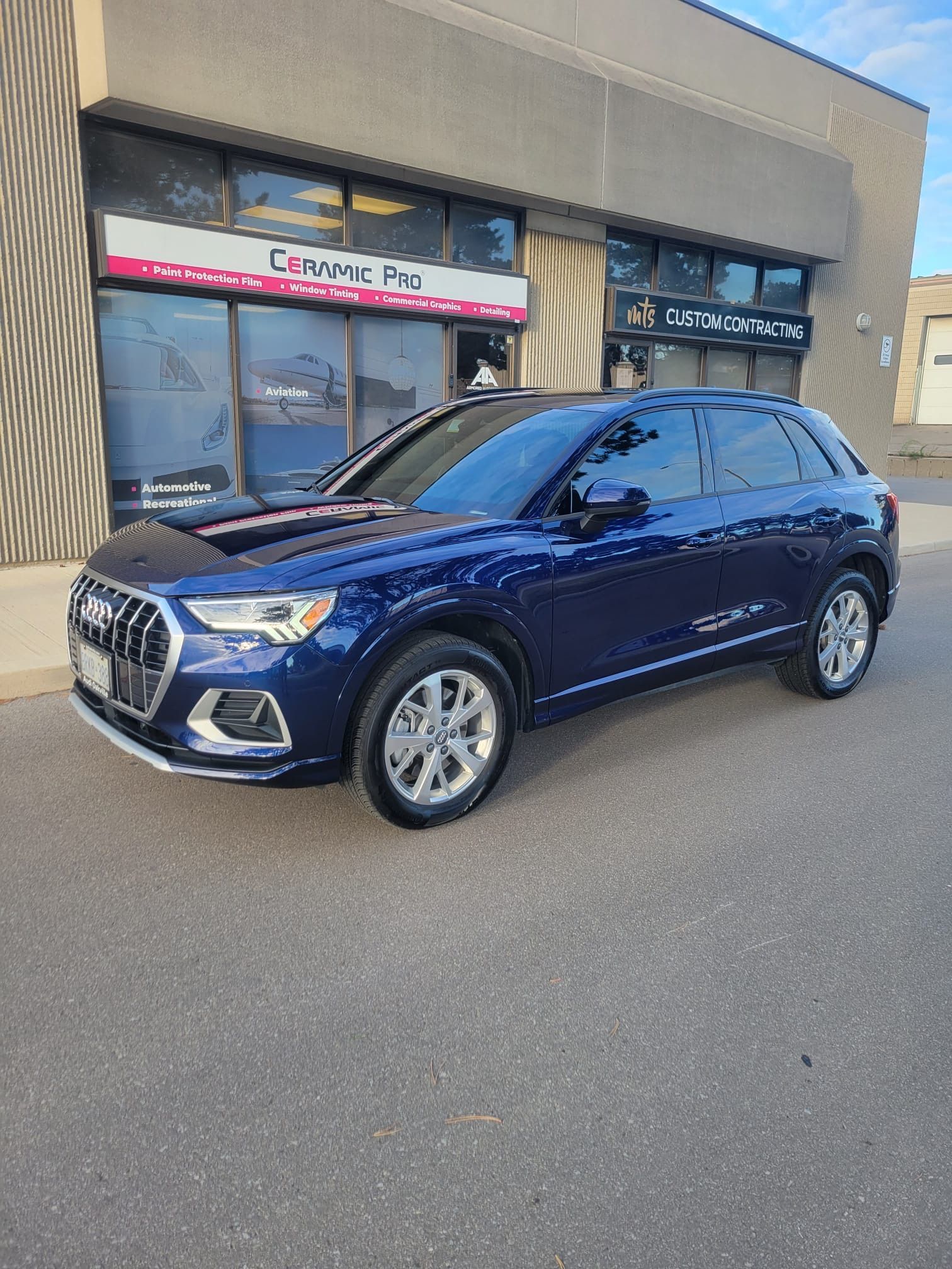 A blue Audi Q3 SUV parked in front of a commercial storefront with large glass windows.