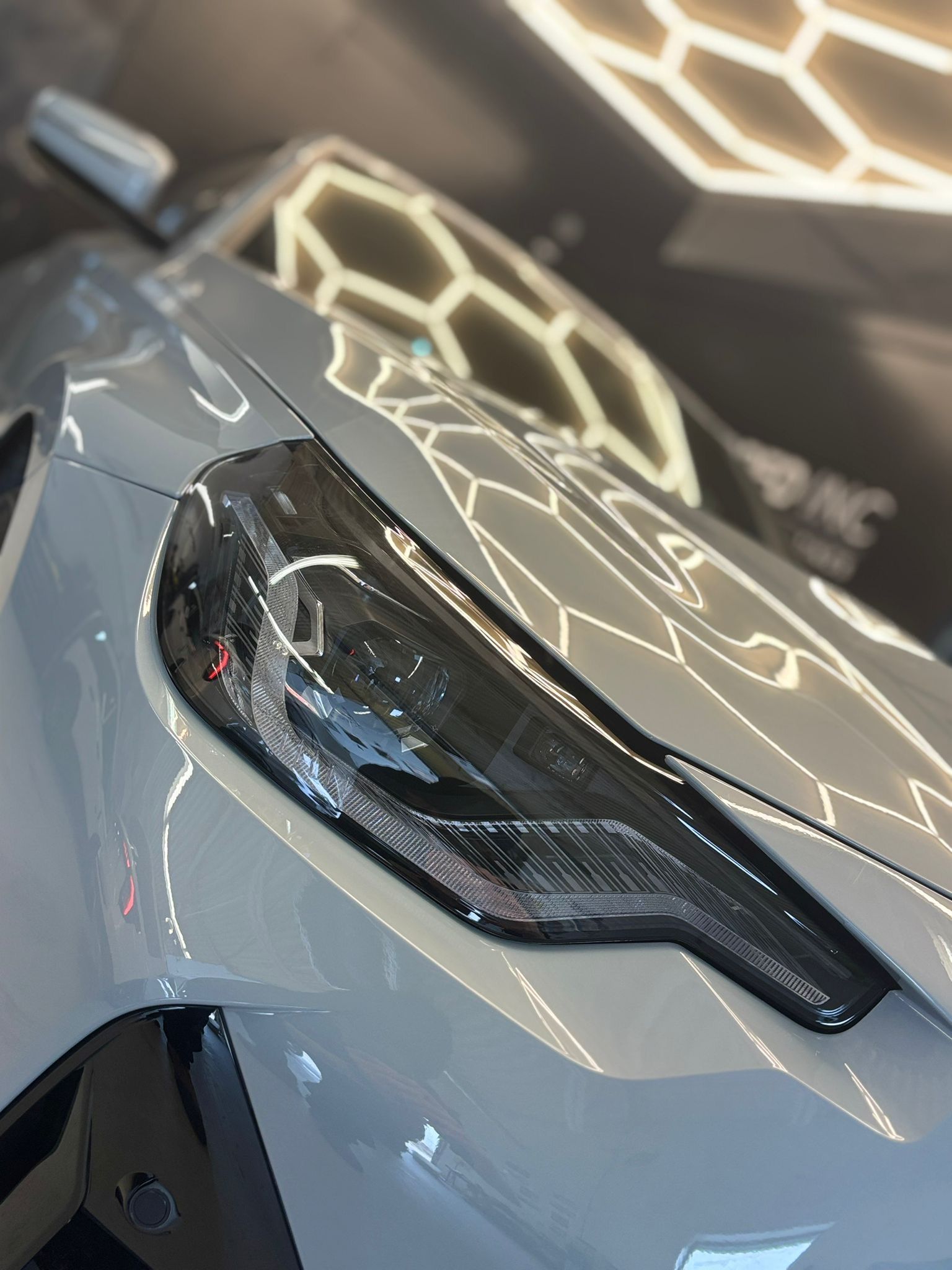 A close-up of a glossy, light grey sports car hood and its sleek, black-tinted LED headlight in a showroom.