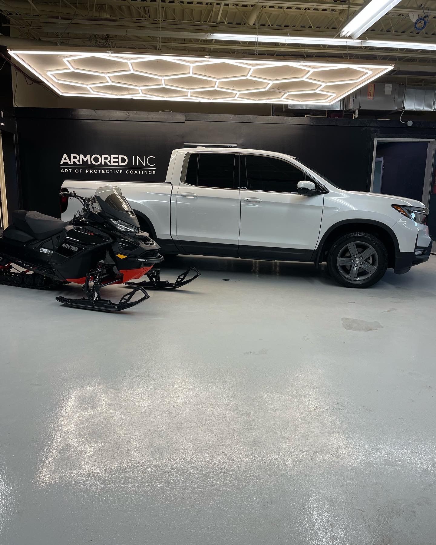 A white pickup truck and a black snowmobile parked in a garage with hexagonal ceiling lights and a black wall sign.