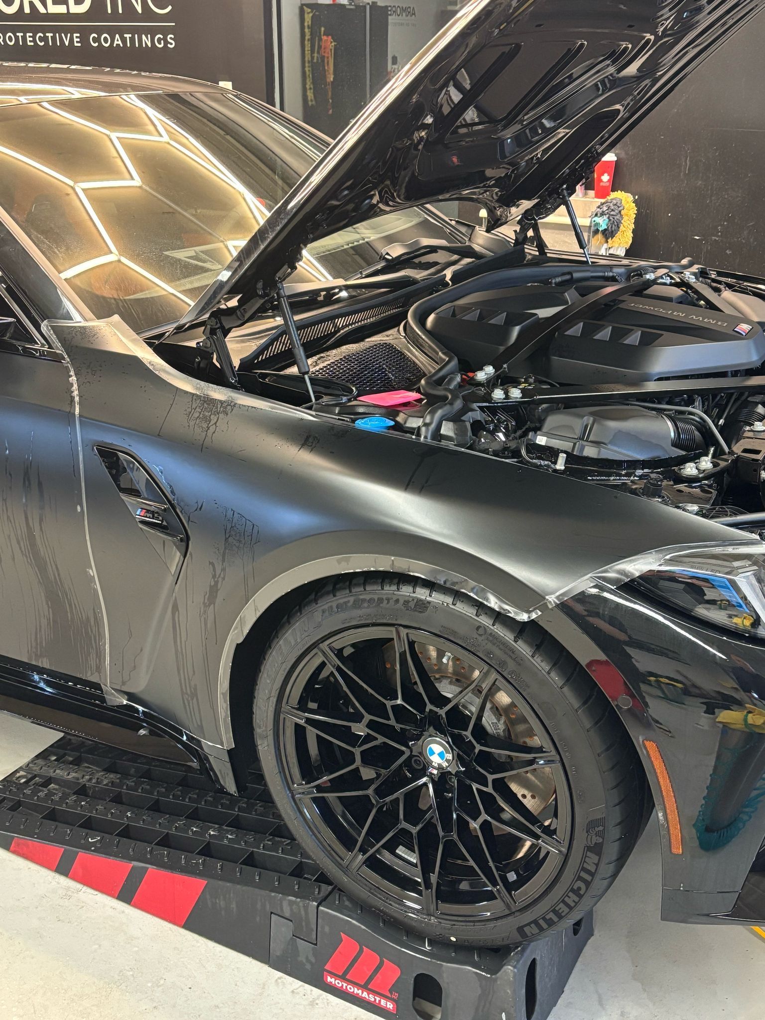 Matte black BMW M4 with hood open, parked on black service ramps in a brightly lit professional detailing garage.