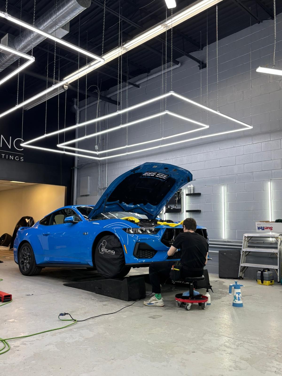 A person working on the engine of a bright blue sports car elevated on ramps in a professional auto detailing shop.