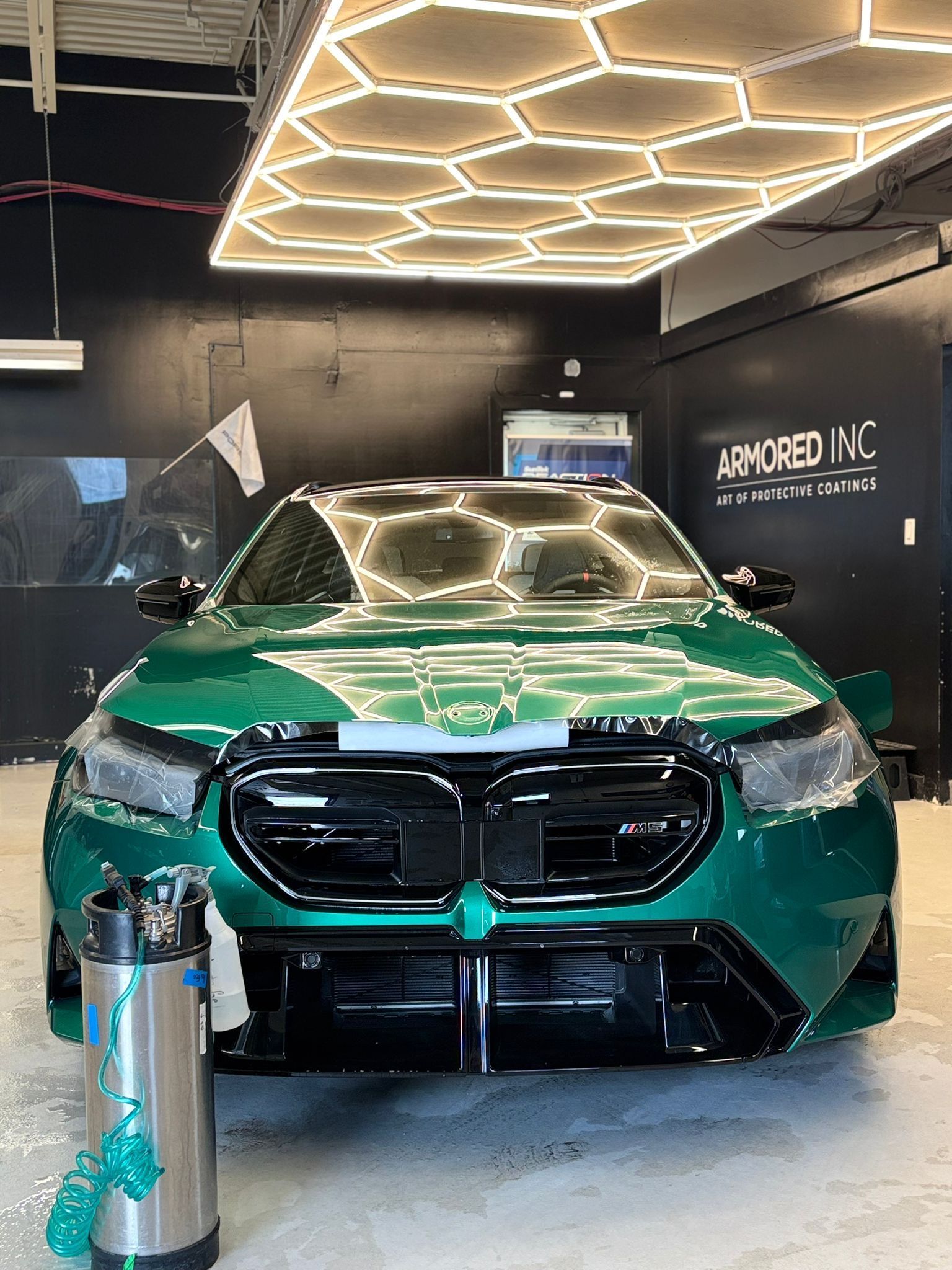 A metallic green BMW parked in a workshop under hexagonal ceiling lights, with a tall silver pressurized tank in front.