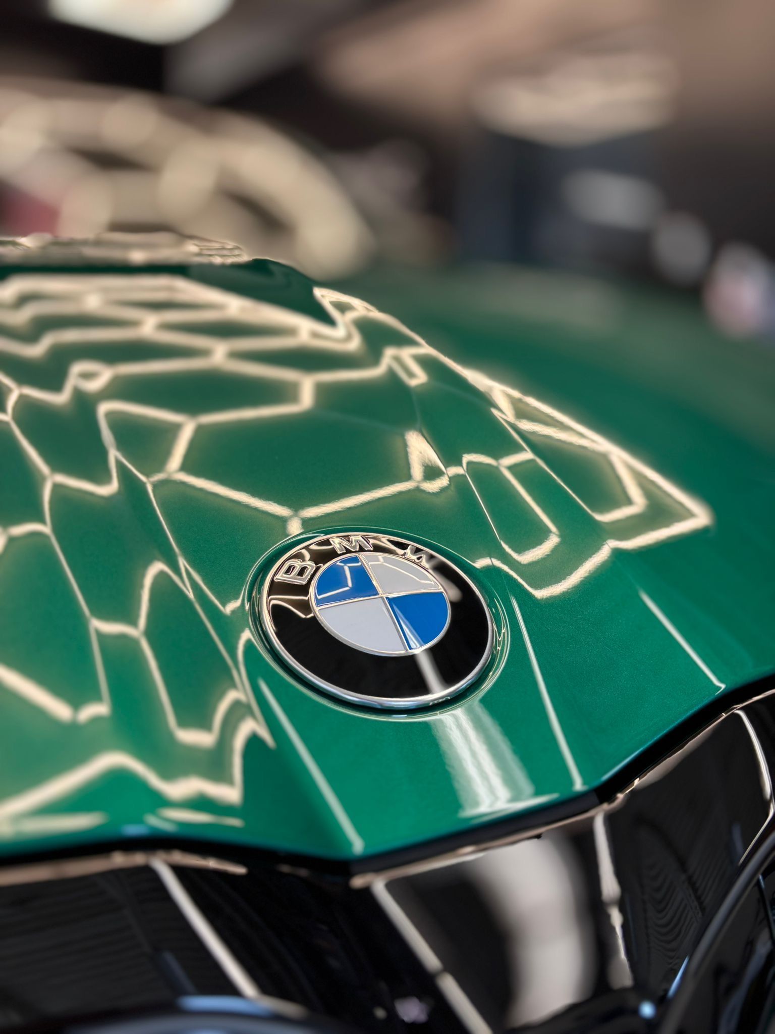 Close-up of a BMW emblem on a metallic green car hood, reflecting hexagonal ceiling lights.