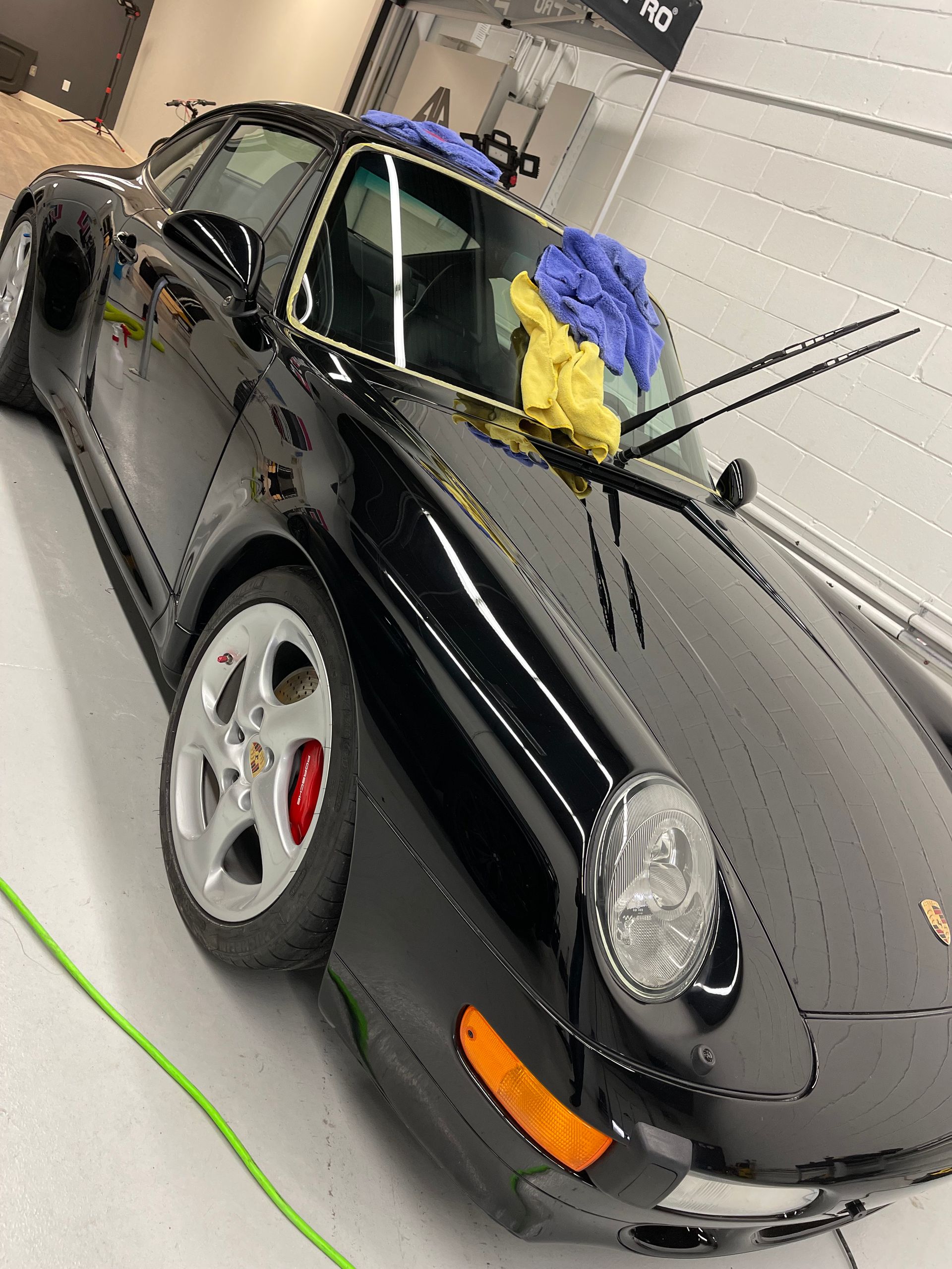 A shiny black Porsche 993 parked in a garage, with blue and yellow detailing cloths resting on the windshield.