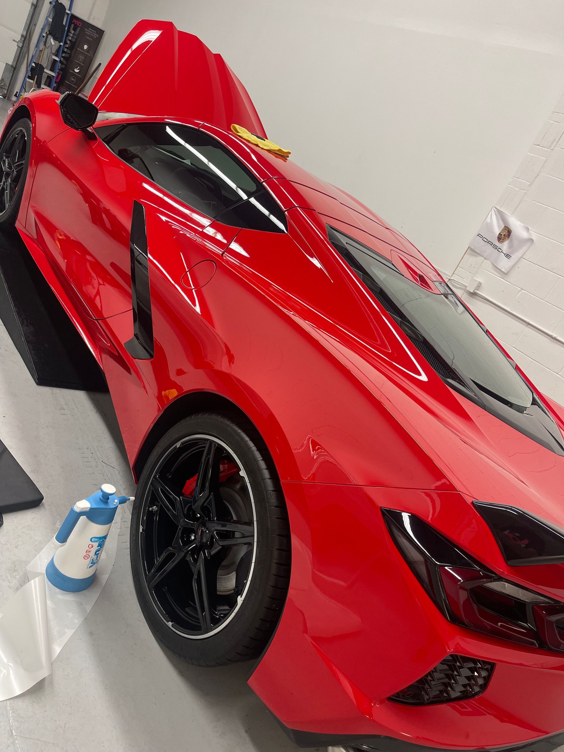 A bright red Corvette sports car parked indoors with a spray bottle nearby and its engine hood partially open.
