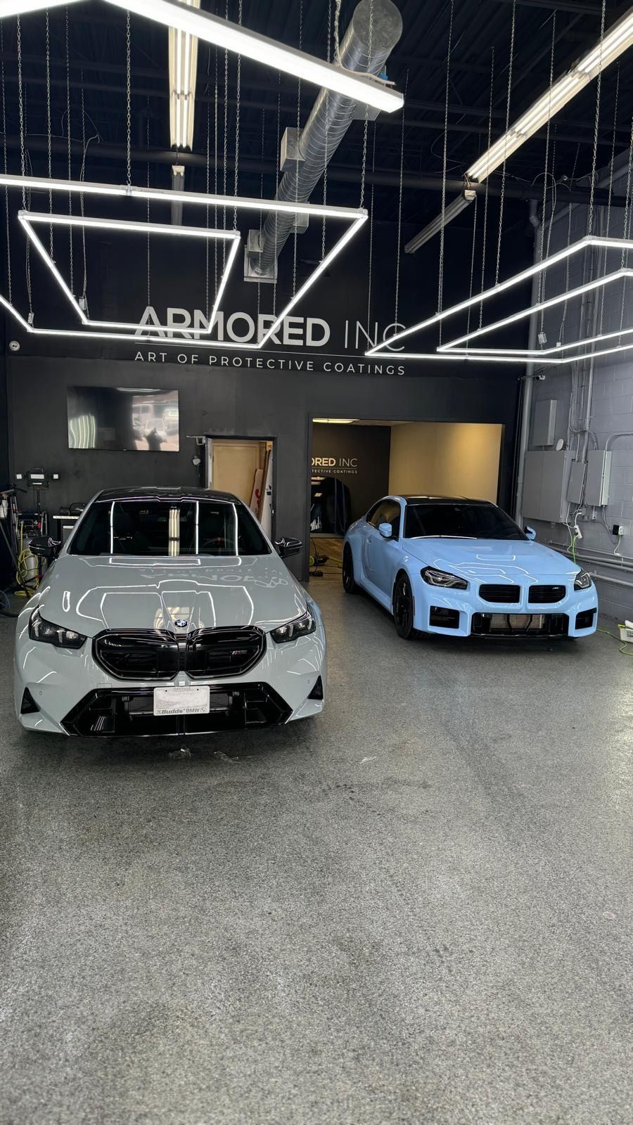 Two luxury BMW cars, one grey and one light blue, parked inside a modern automotive studio with bright square lighting.