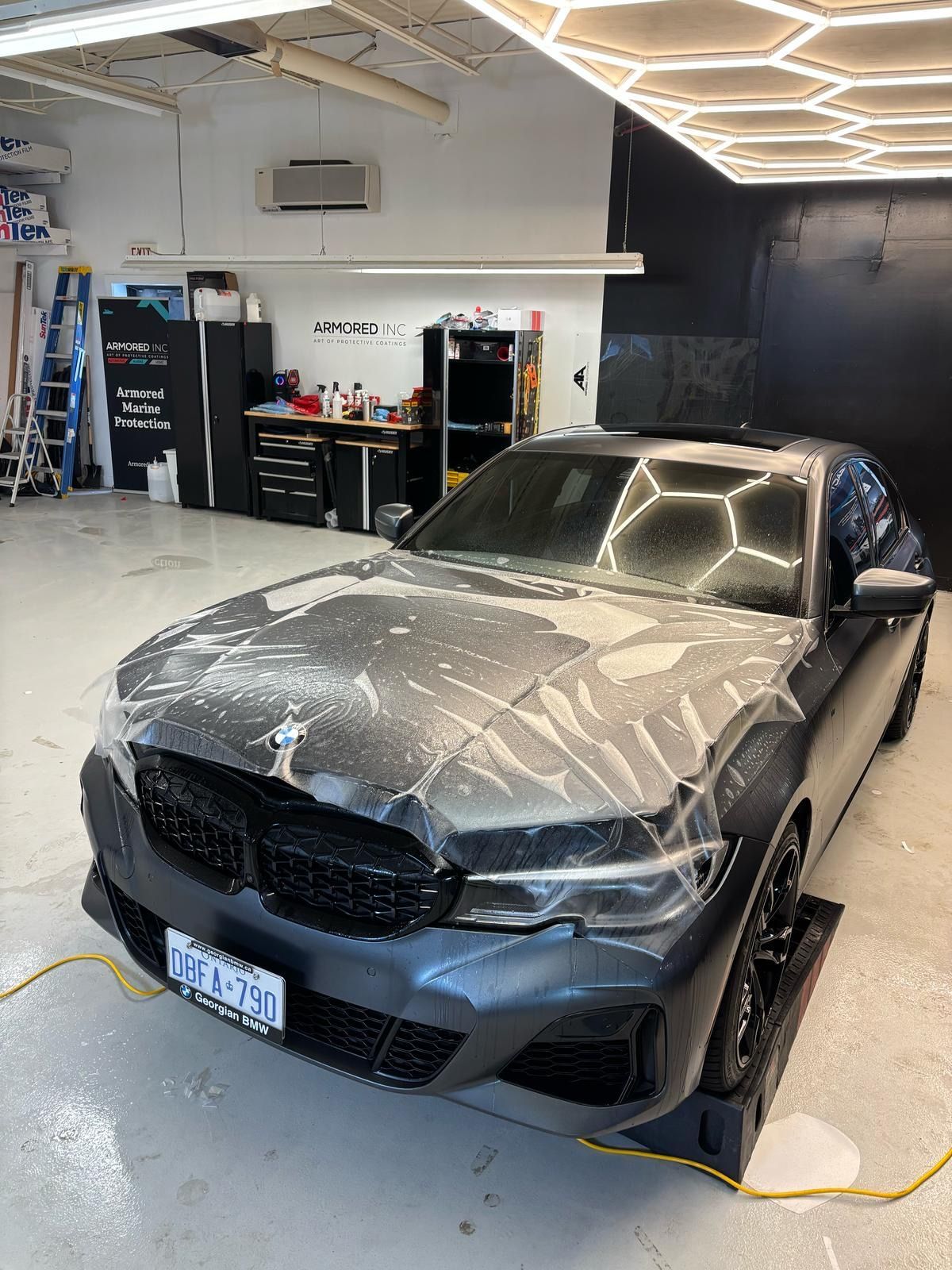 A matte grey BMW parked in a workshop undergoing the application of a transparent paint protection film on its hood.
