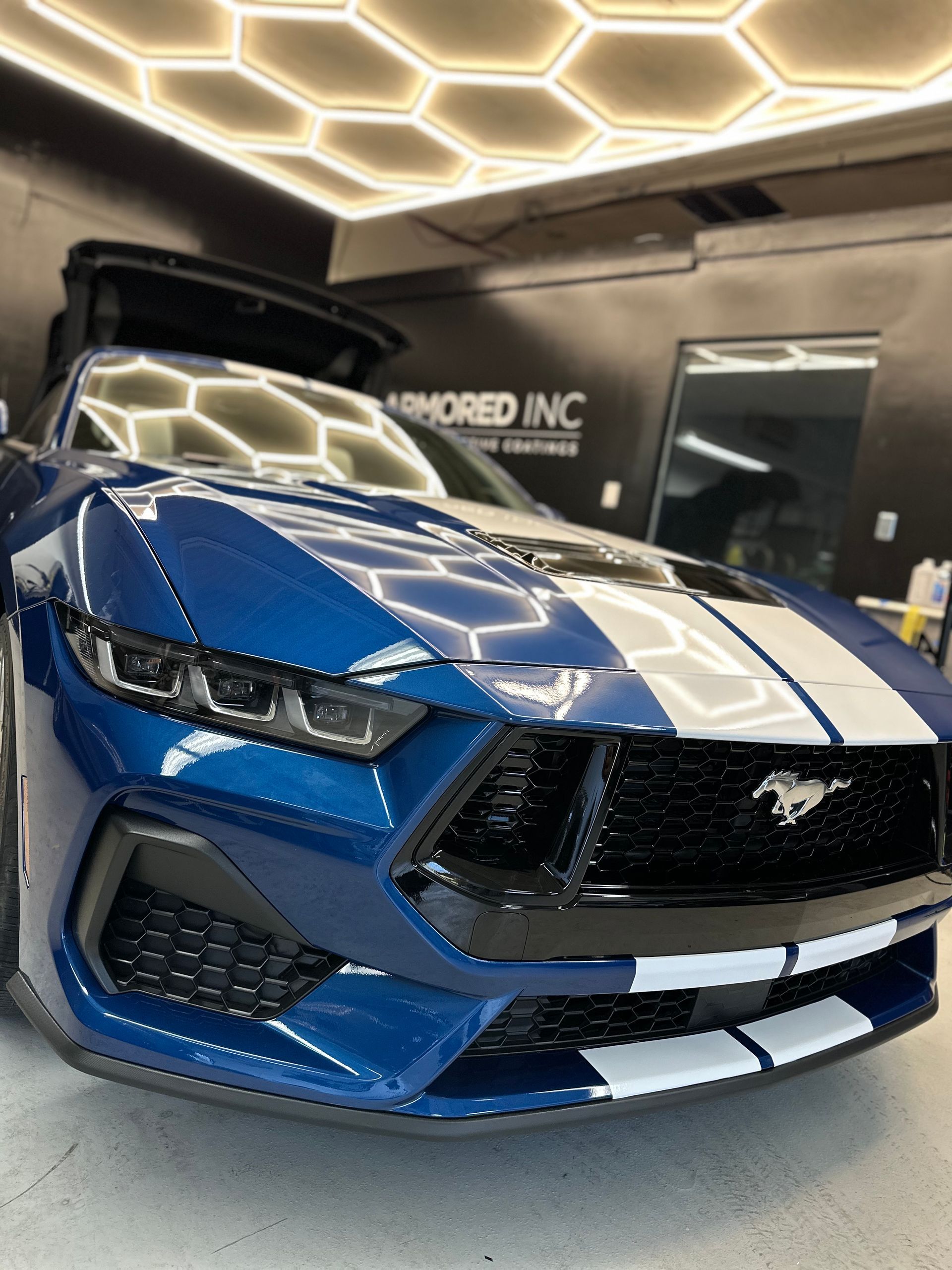 A glossy blue Ford Mustang with white racing stripes parked in a garage with geometric ceiling lights.