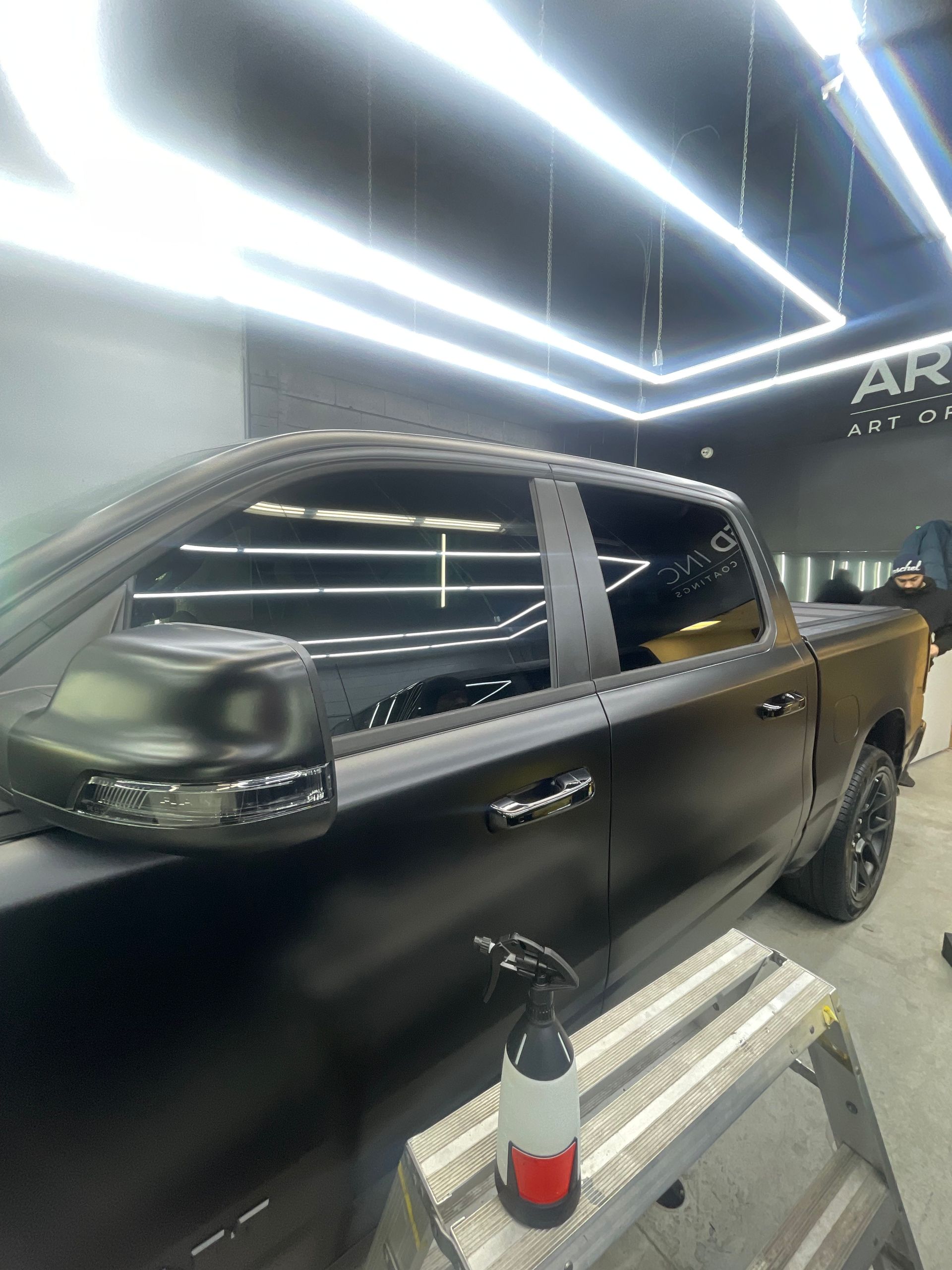 A matte black pickup truck parked in a well-lit workshop with a spray bottle on a step ladder in the foreground.