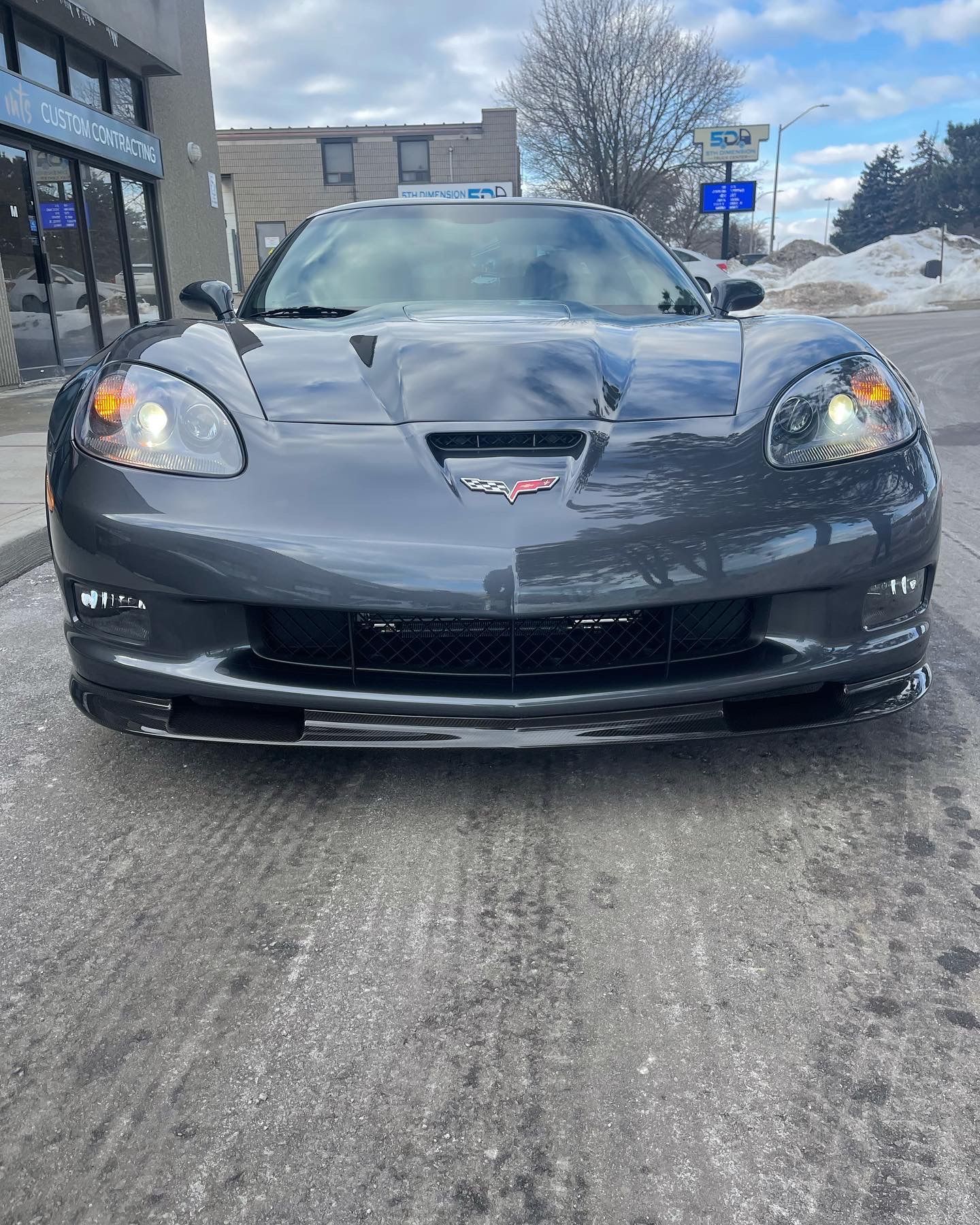 A grey Chevrolet Corvette sports car parked on a paved street with a building and snowy landscape in the background.
