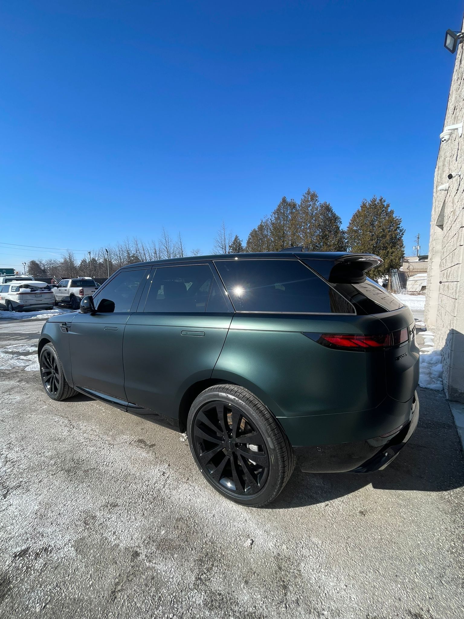 A dark matte green Range Rover Sport parked on a gravel lot under a clear blue sky, seen from the rear three-quarter view.