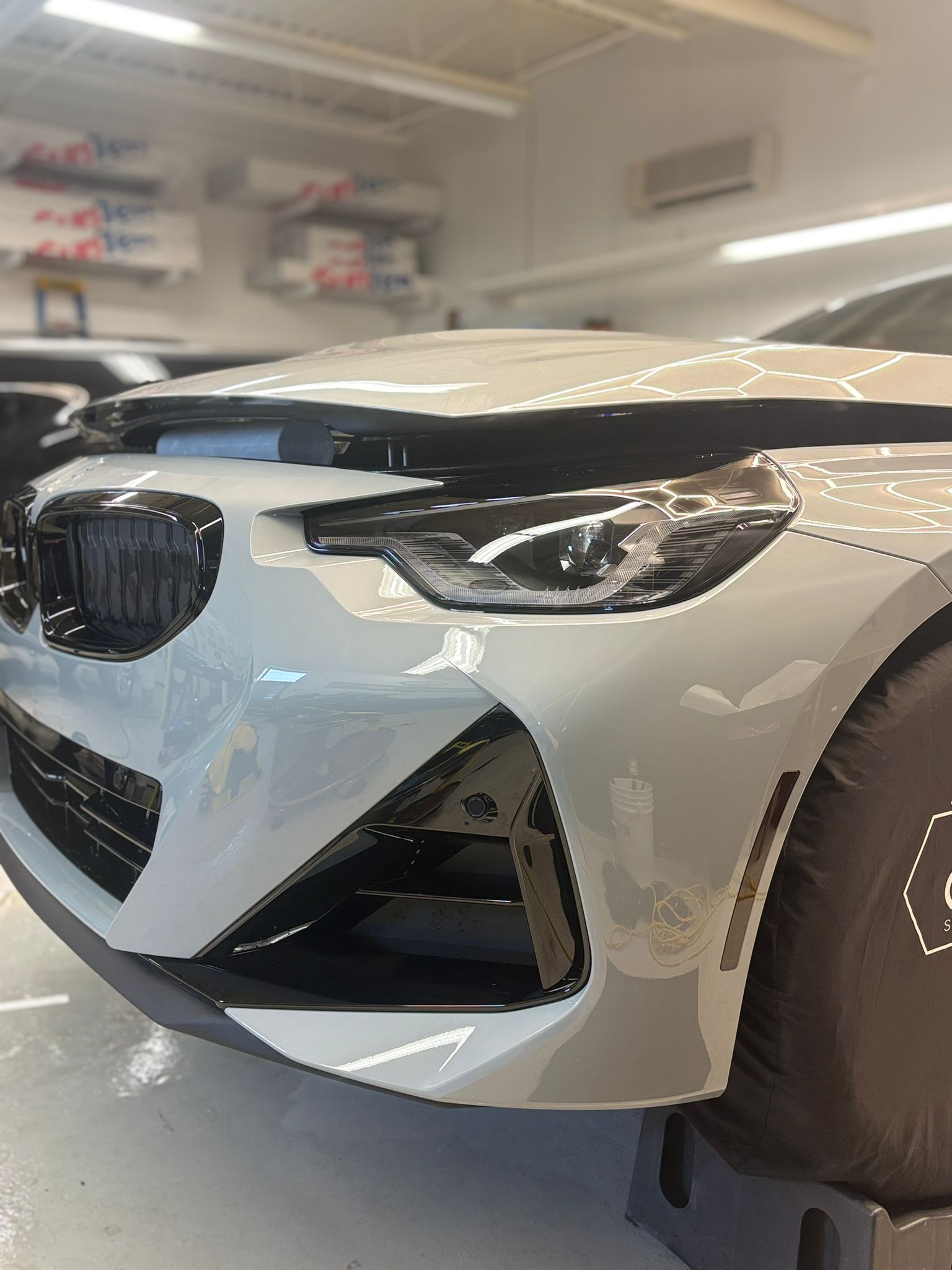 Close-up of a light gray BMW car’s front end, including the headlight, grille, and bumper, inside an auto shop.
