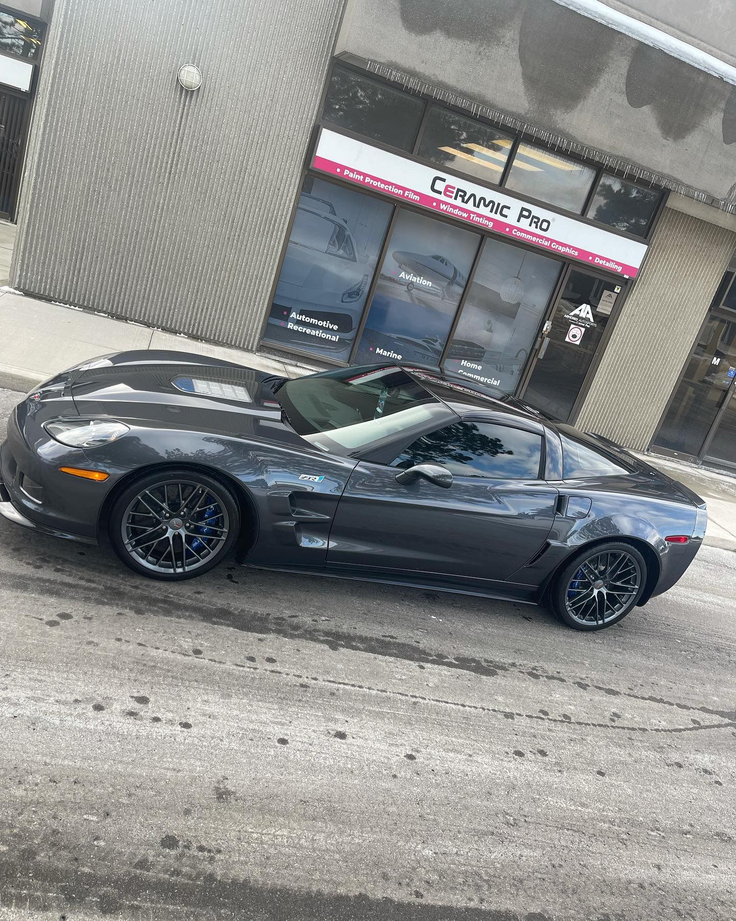 A dark gray Chevrolet Corvette parked in front of a Ceramic Pro automotive shop.