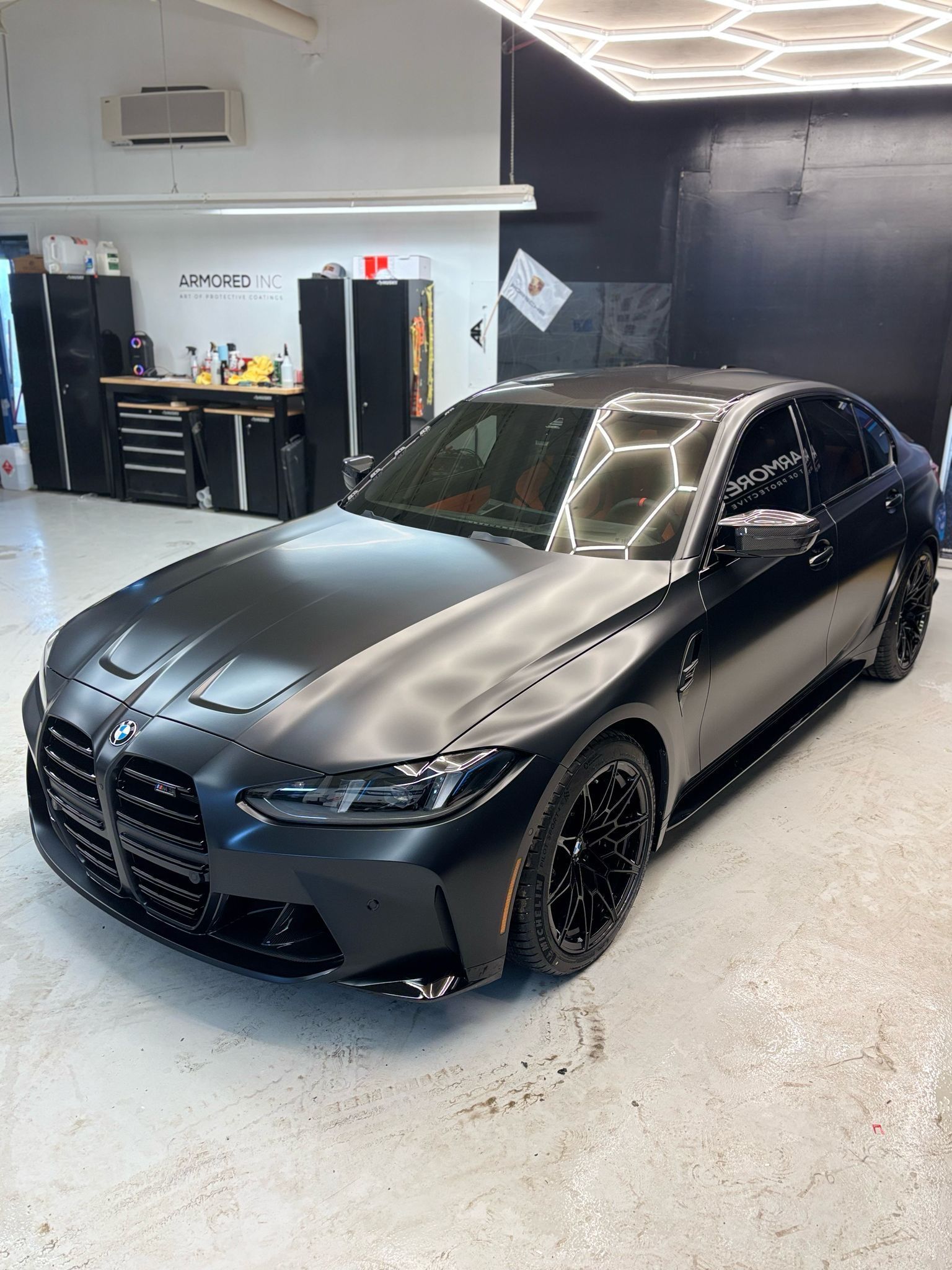 A matte black BMW M3 sports sedan parked inside a brightly lit professional car detailing shop.