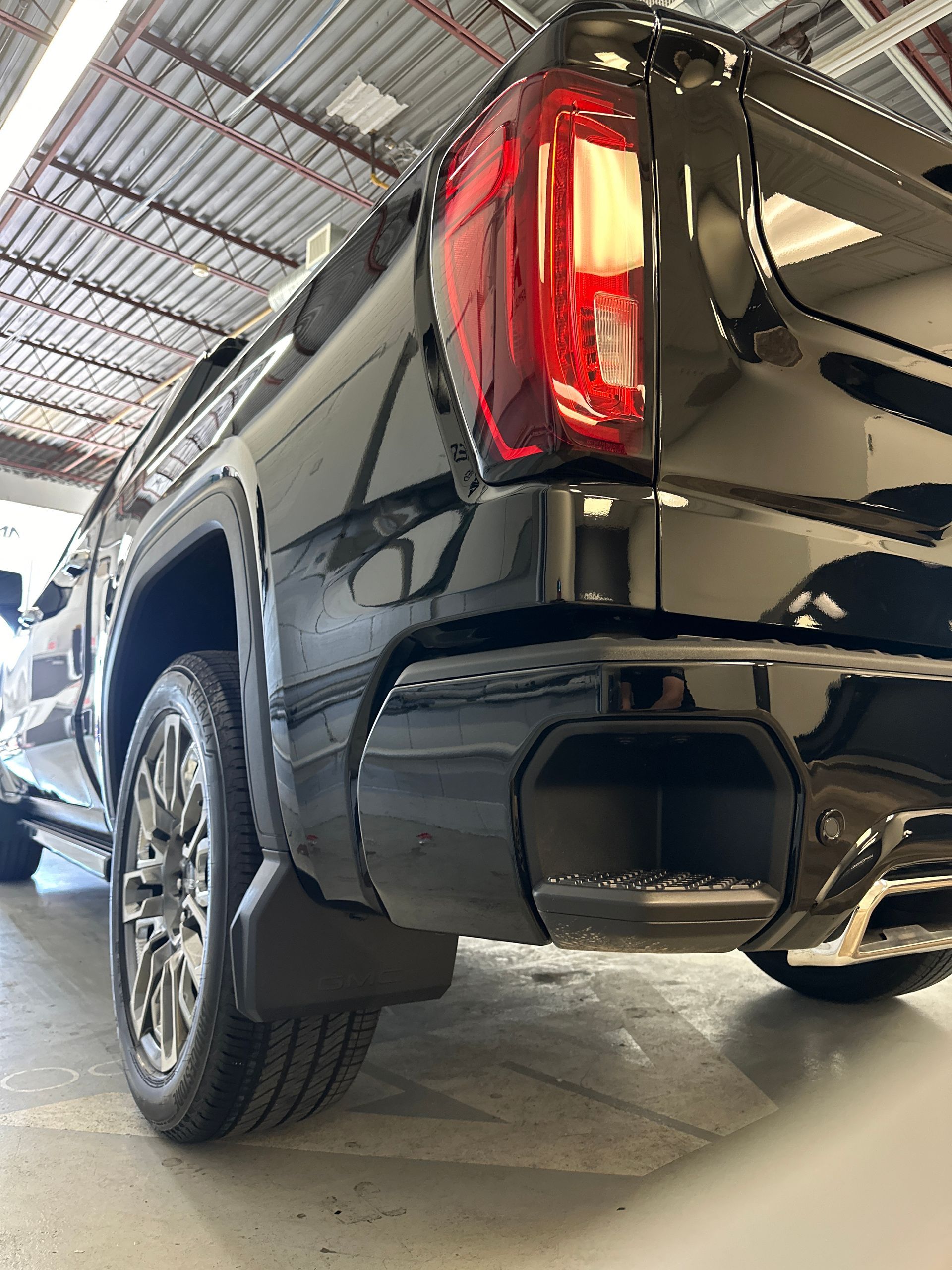 A low-angle view of the rear of a shiny black GMC pickup truck, showing the tail light, bumper step, and rear tire.