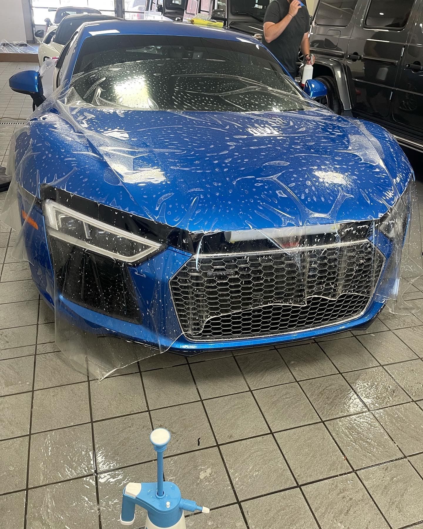 A blue Audi R8 in a garage being covered with a transparent protective film, with a spray bottle in the foreground.