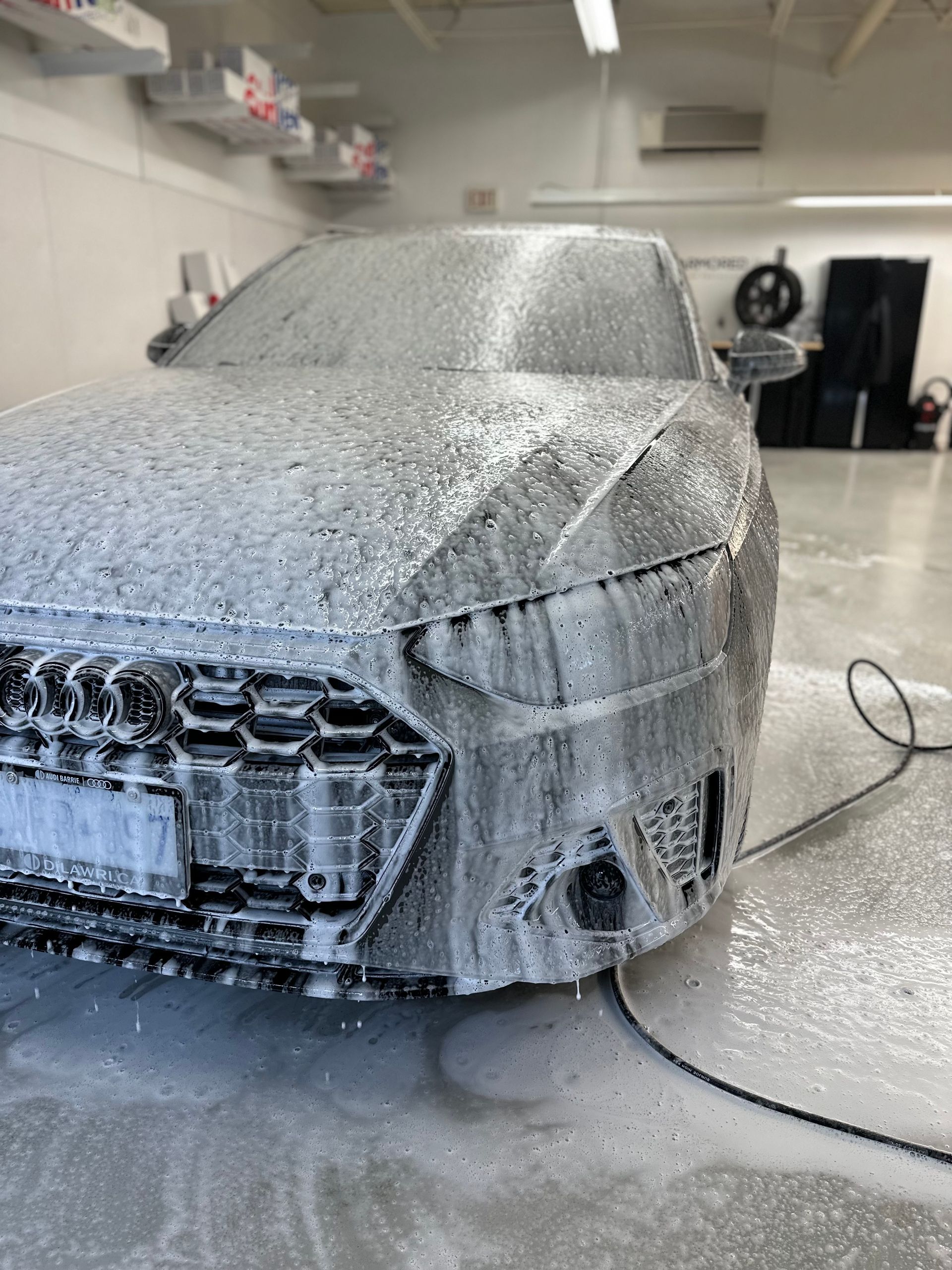 A car covered in thick white soap suds in a brightly lit garage, during a professional cleaning service.