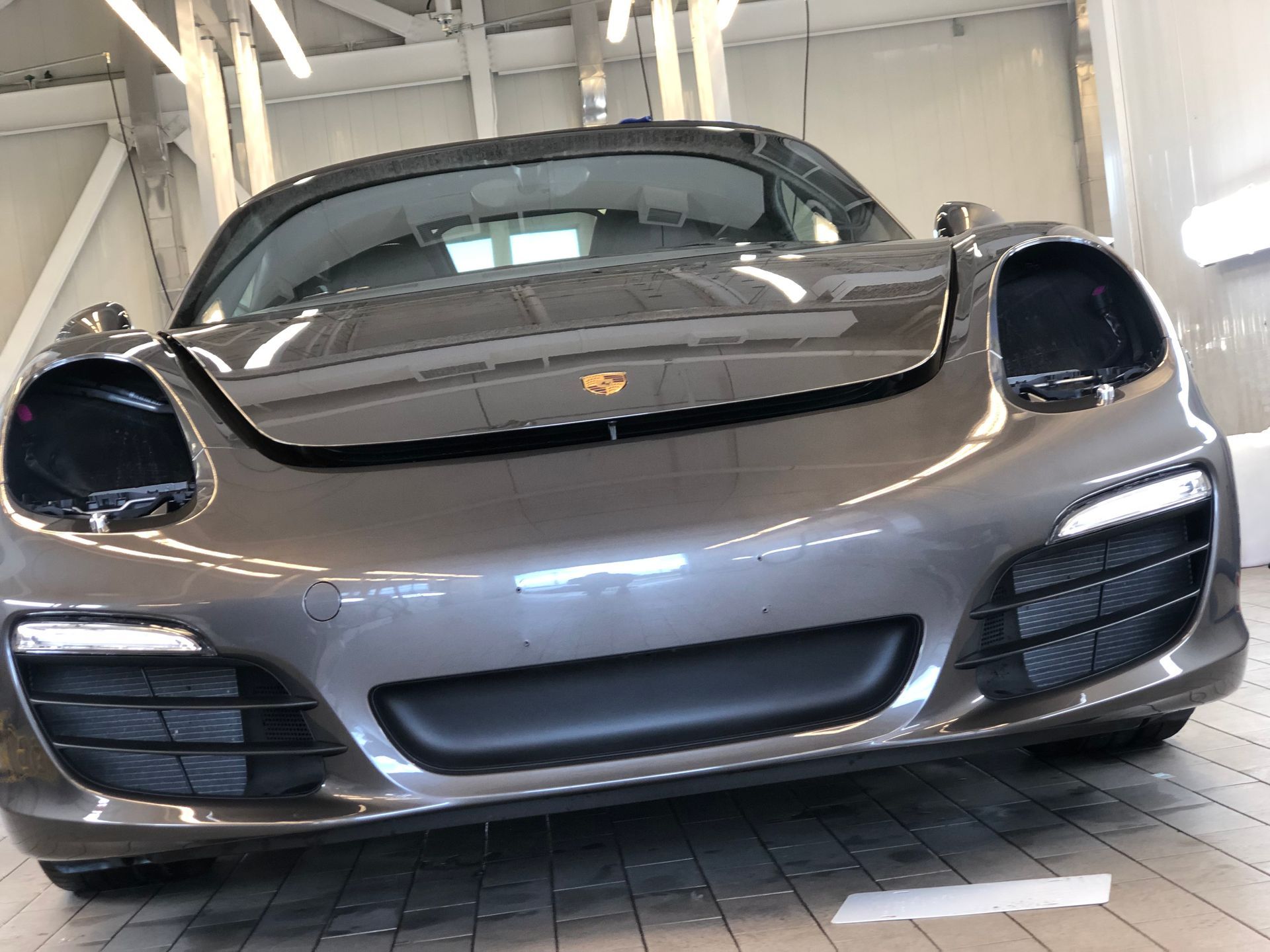 A dark gray Porsche sports car sits in a brightly lit garage, missing its main headlights.