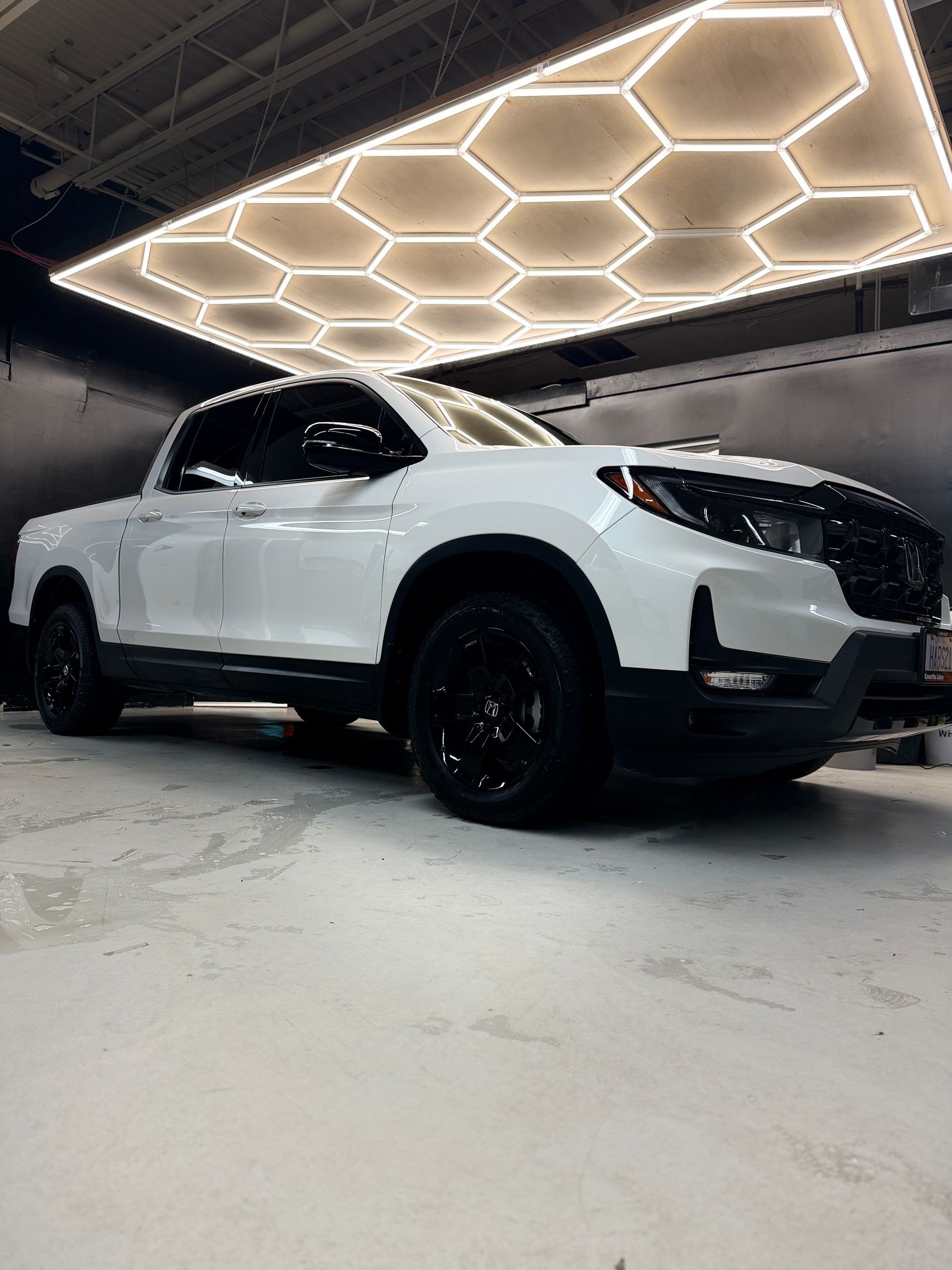A white Honda Ridgeline truck with black rims parked in a garage under hexagonal LED lighting.