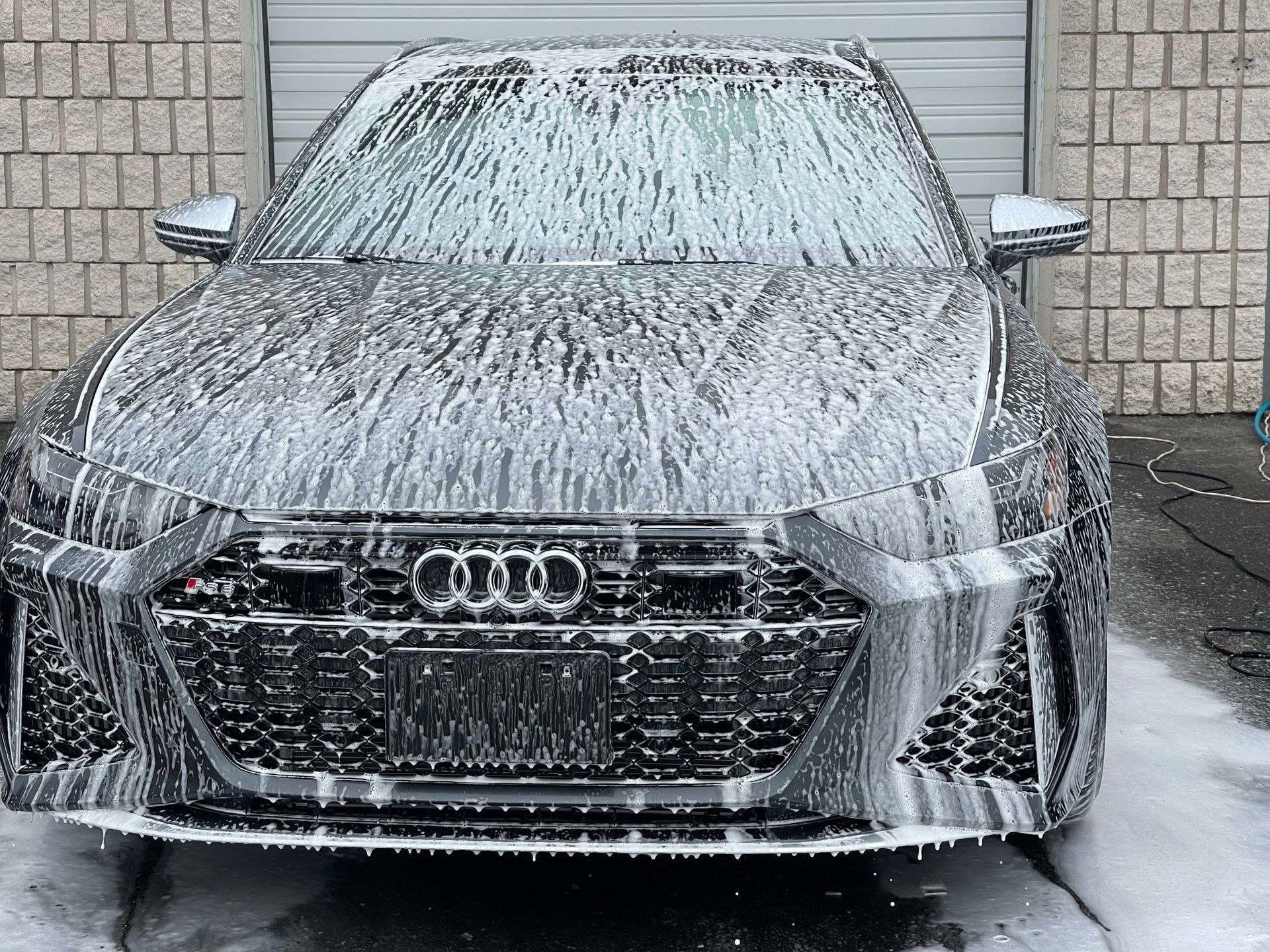An Audi sedan covered in thick white soap suds during a car wash, parked in front of a brick wall.