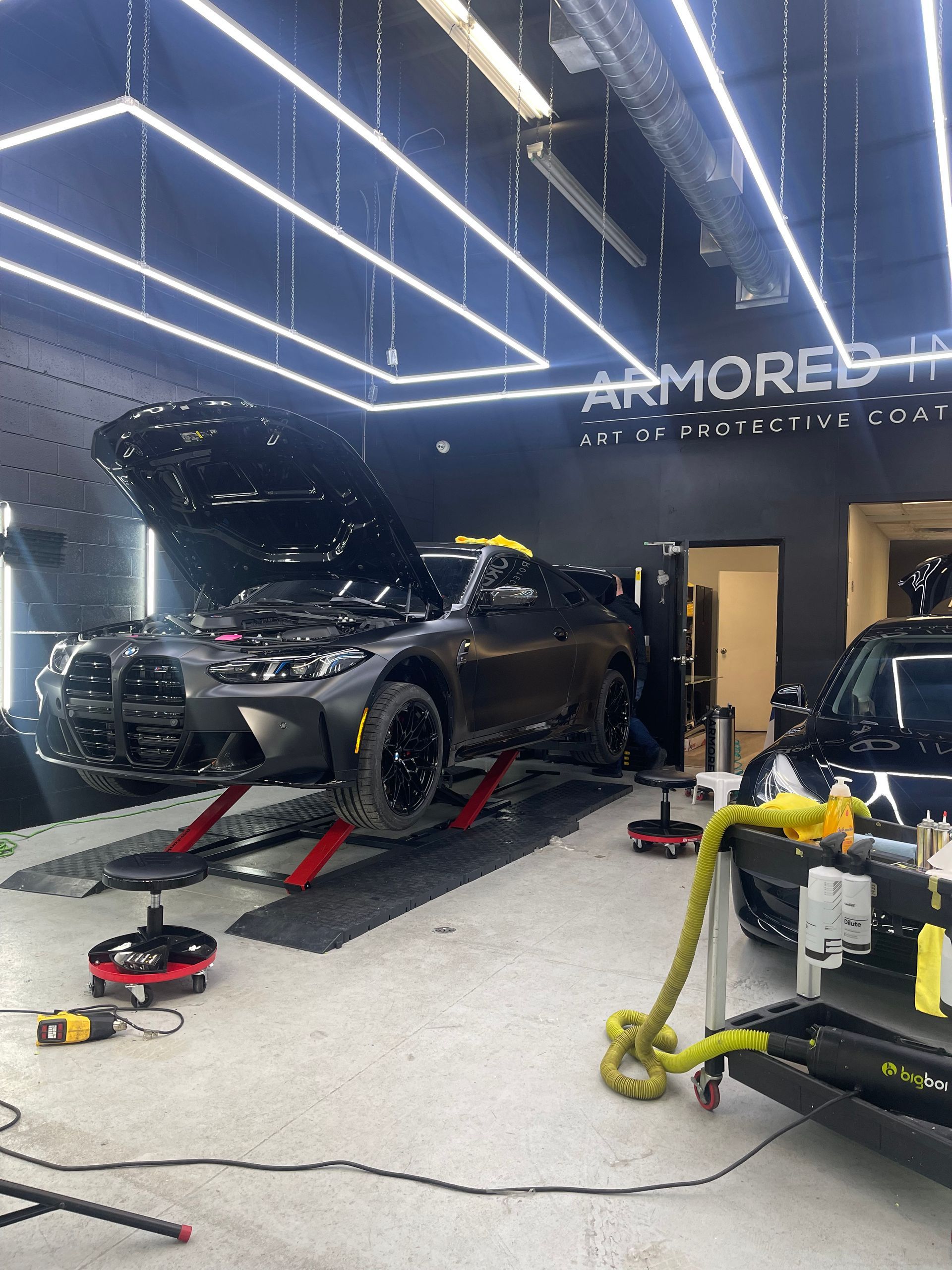 A grey car with its hood open sits on a lift in a brightly lit automotive workshop with another car parked nearby.