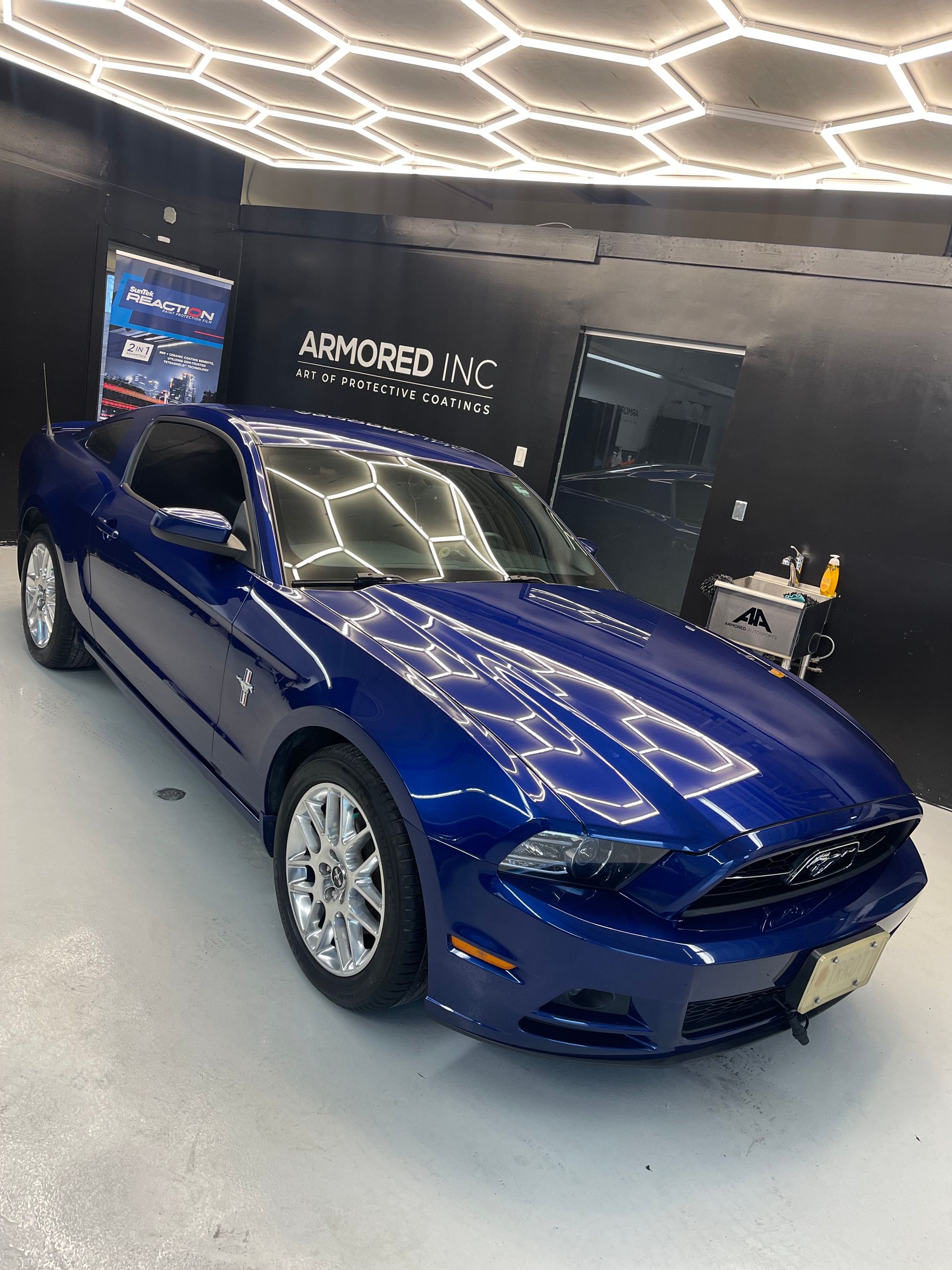 A glossy blue Ford Mustang parked inside a garage with patterned ceiling lights and a black wall sign reading Armored NYC.