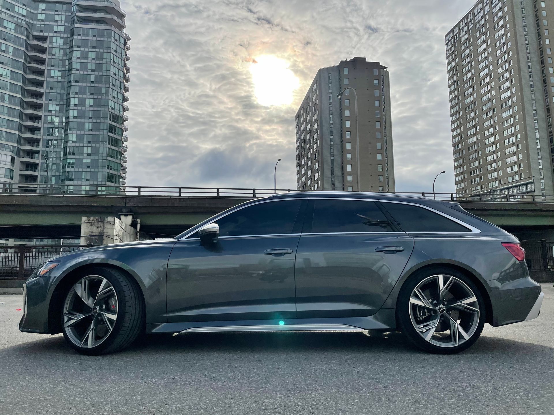 A dark gray Audi RS6 Avant wagon parked on a paved lot with high-rise buildings and a bridge in the background.
