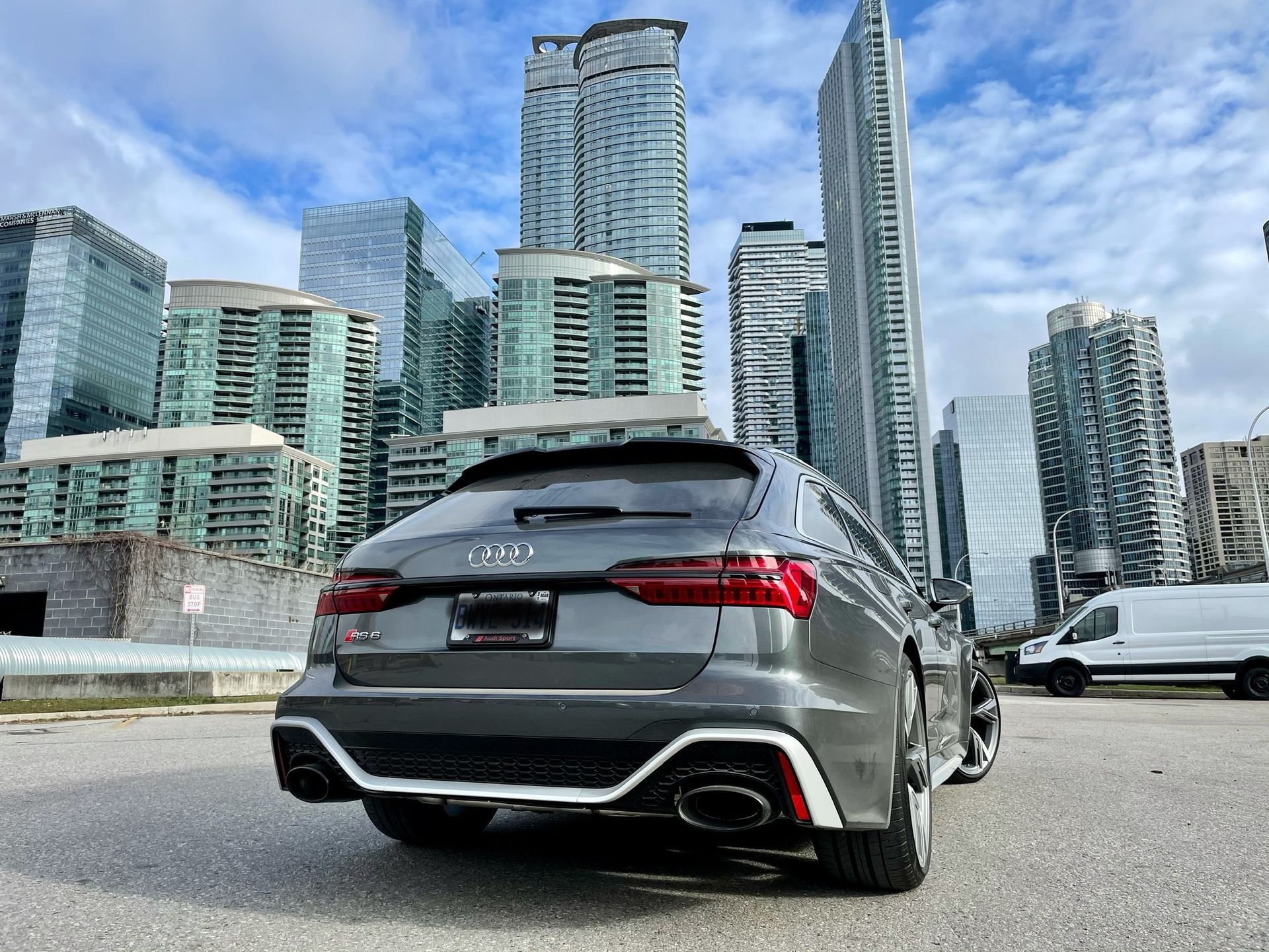 A gray Audi station wagon parked on an urban lot against a backdrop of towering modern glass skyscrapers under a blue sky.